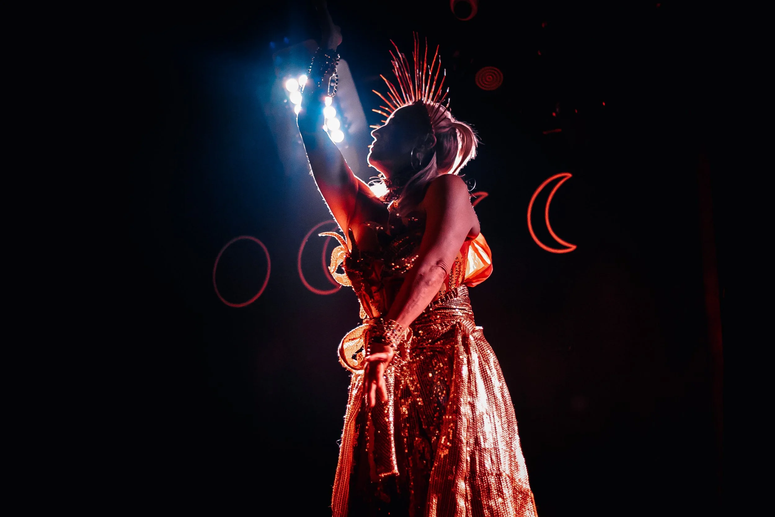 Performer dressed in shiny red outfit with a spiked crown, backlit on stage with neon symbols in the background.
