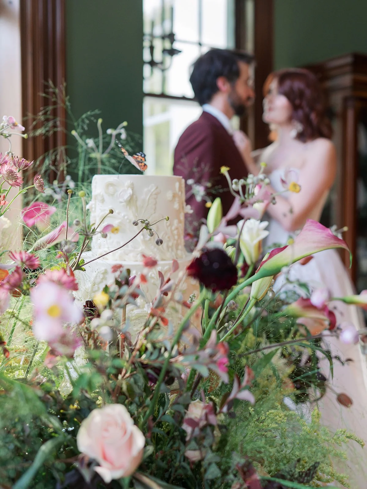 A wedding cake on a table surrounded by a lush arrangement of pink, white, and dark-colored flowers, with a blurred background of a bride and groom.