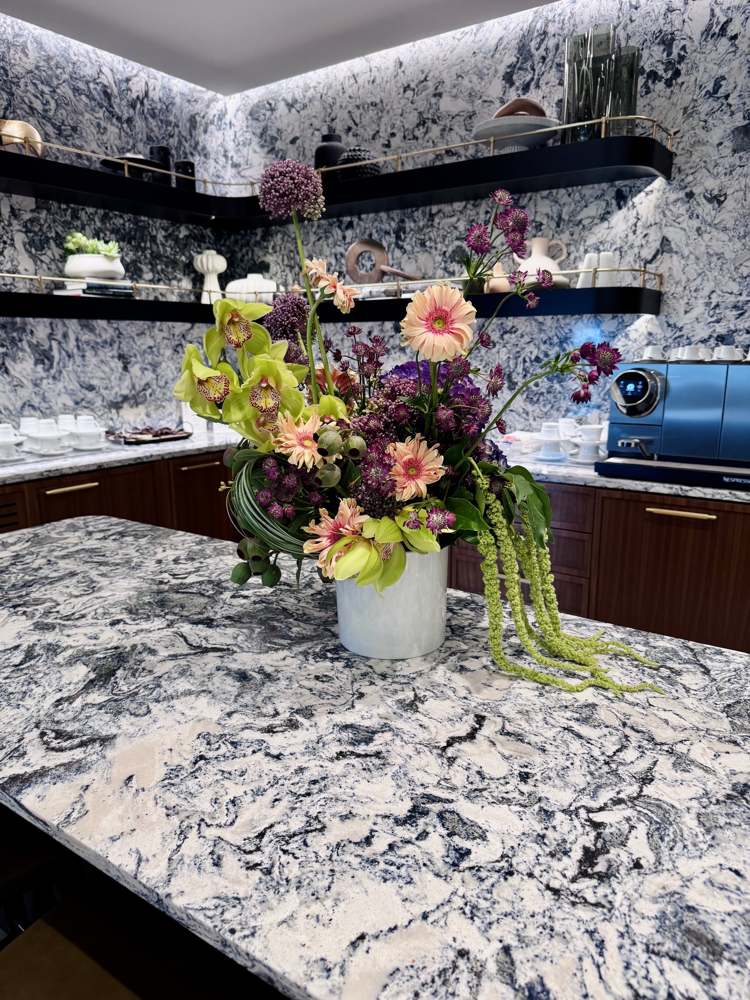 A floral arrangement in a white vase on a kitchen countertop with a blue and white marbled pattern. The background features two black shelves with decorative items and a blue coffee machine.