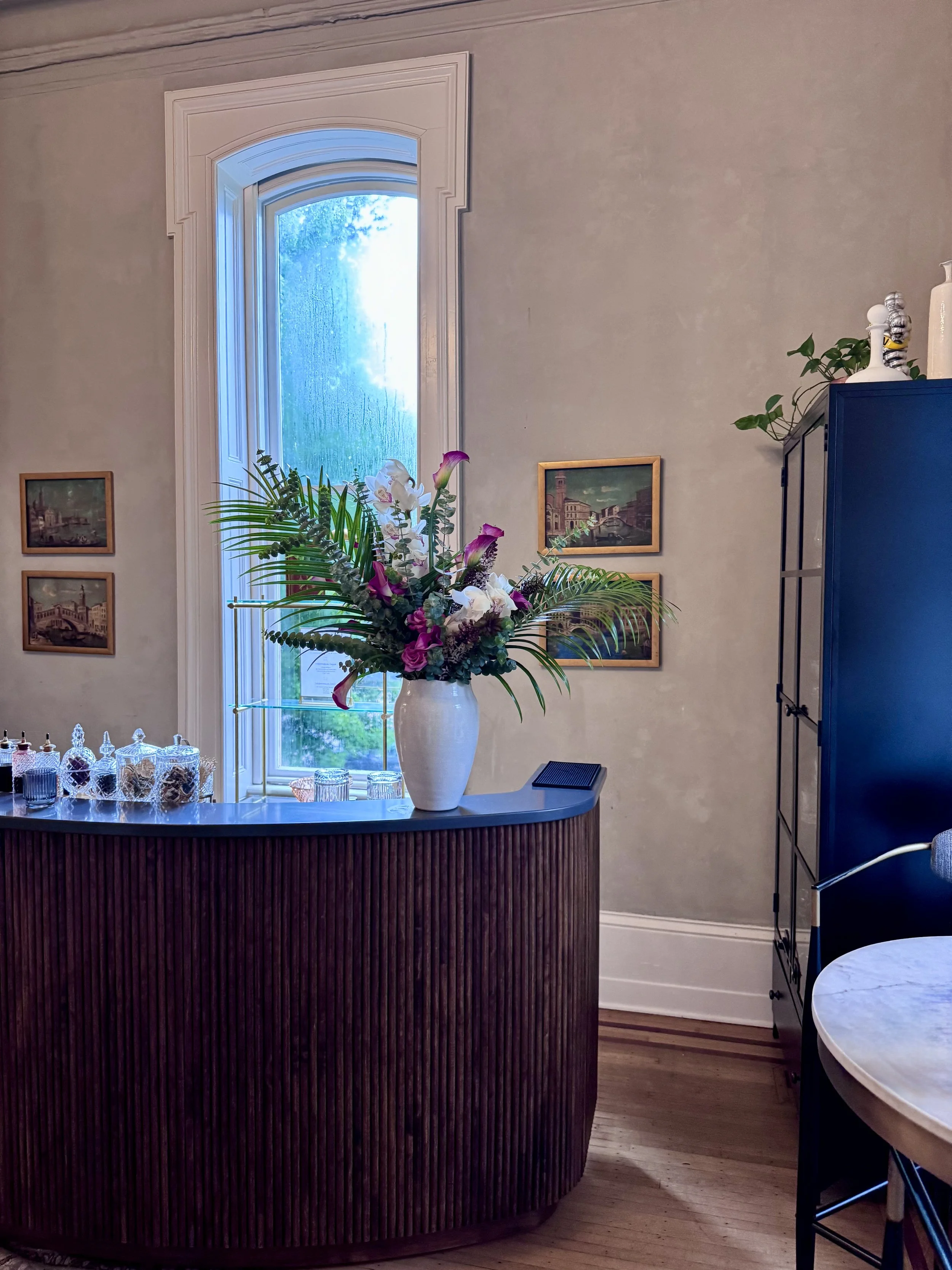A cozy interior with a decorated bar counter, a large vase of flowers, a window showing rain, framed pictures on the wall, and a cabinet with decorative items.
