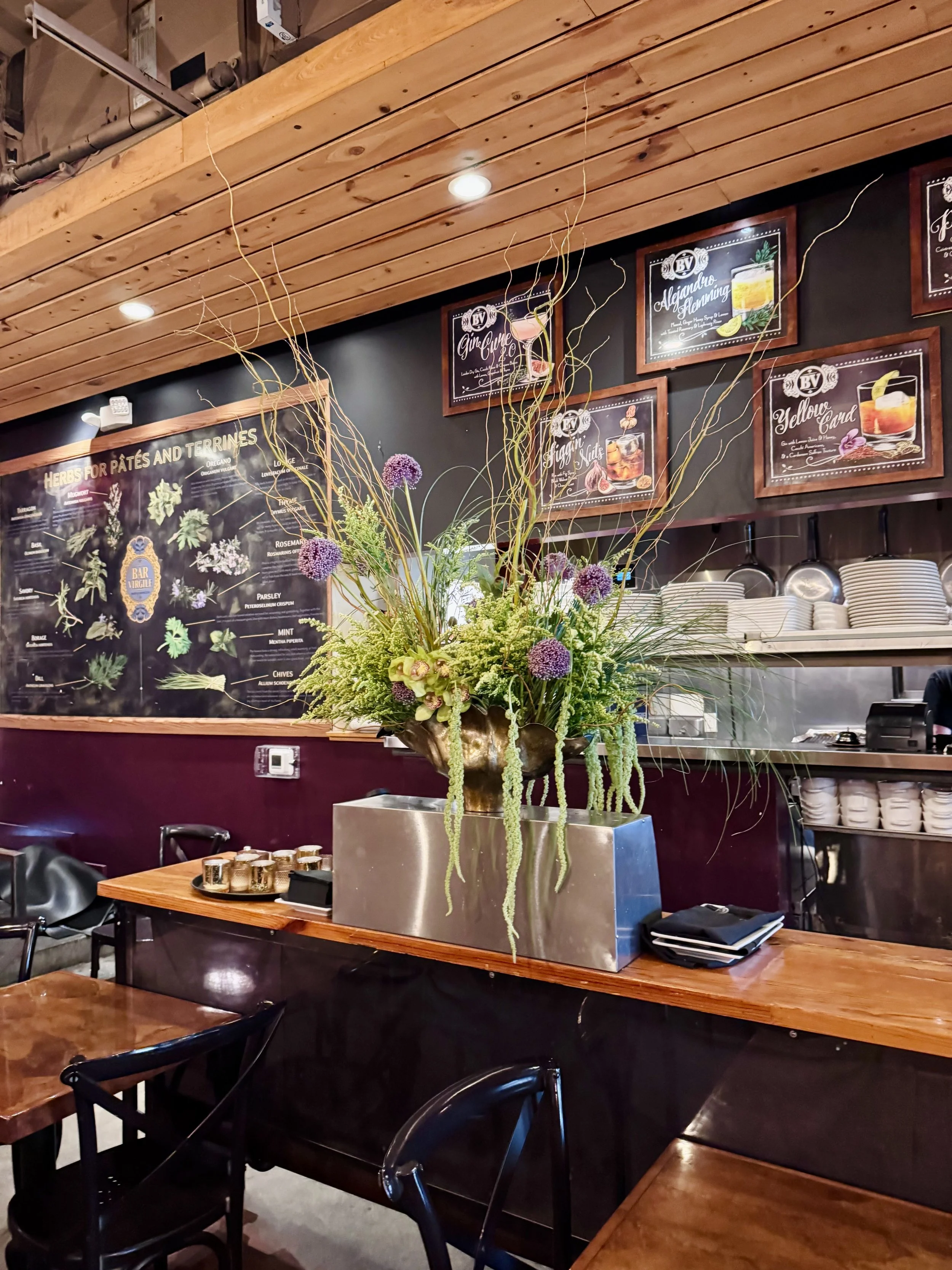 Interior of a restaurant or cafe with wooden tables and black chairs. A large floral arrangement with purple flowers and greenery is in a metallic vase on a table. The back wall features a black chalkboard menu with gold and white lettering and a wooden border, displaying herbs for pates and terrines. Above the menu, framed chalkboard-style posters list drink names and images. The ceiling is wooden with embedded lights.
