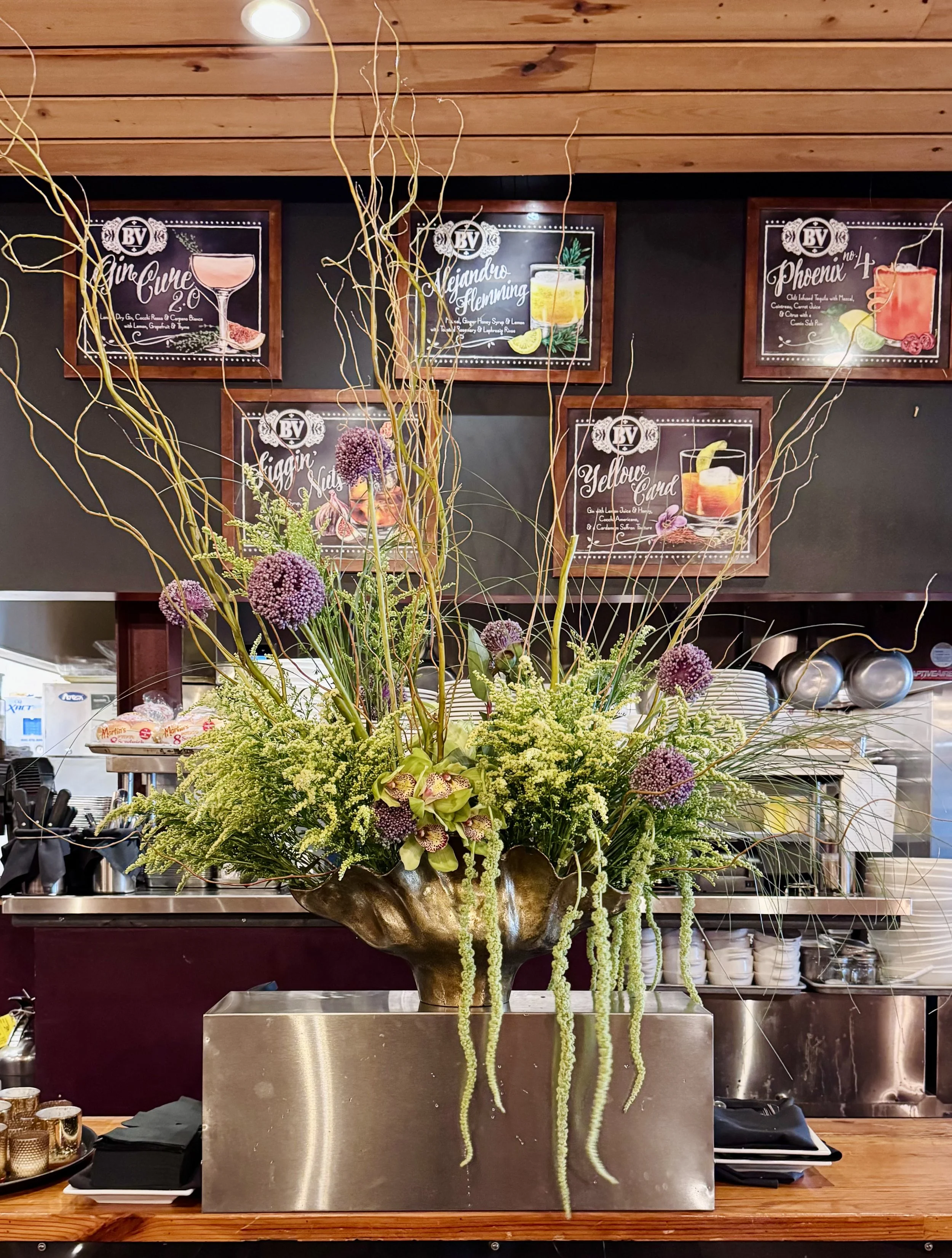 A floral arrangement with purple and green flowers and long green hanging strands in a metallic vase on a wooden counter in front of a chalkboard menu wall with drink options. 