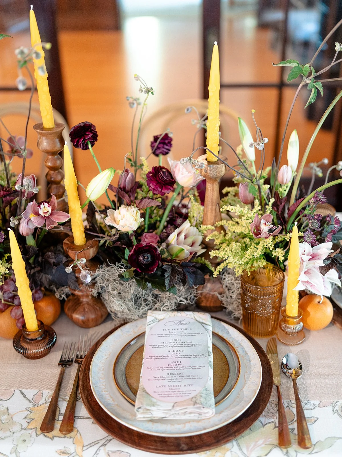 A decorated table with a floral centerpiece, yellow candles, and a place setting with a menu card placed on a plate.