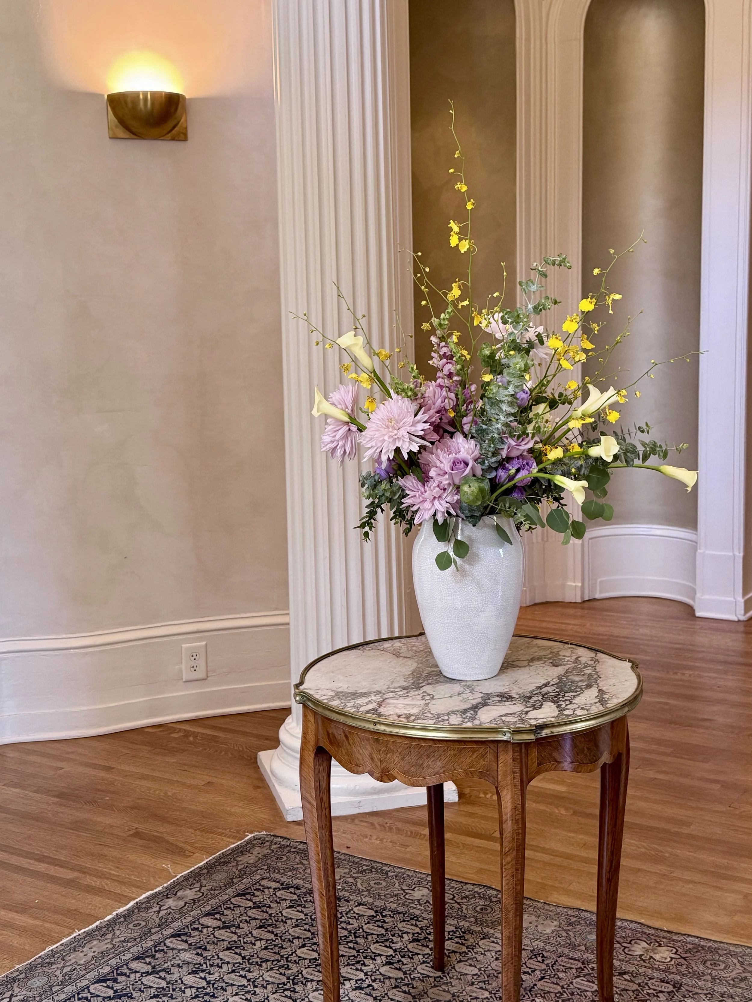 A large white ceramic vase with flowers, including pink dahlias, yellow flowers, and lilies, set on a vintage wooden table with a marble top, in a room with light-colored walls, a floor lamp, crown molding, and wooden flooring.