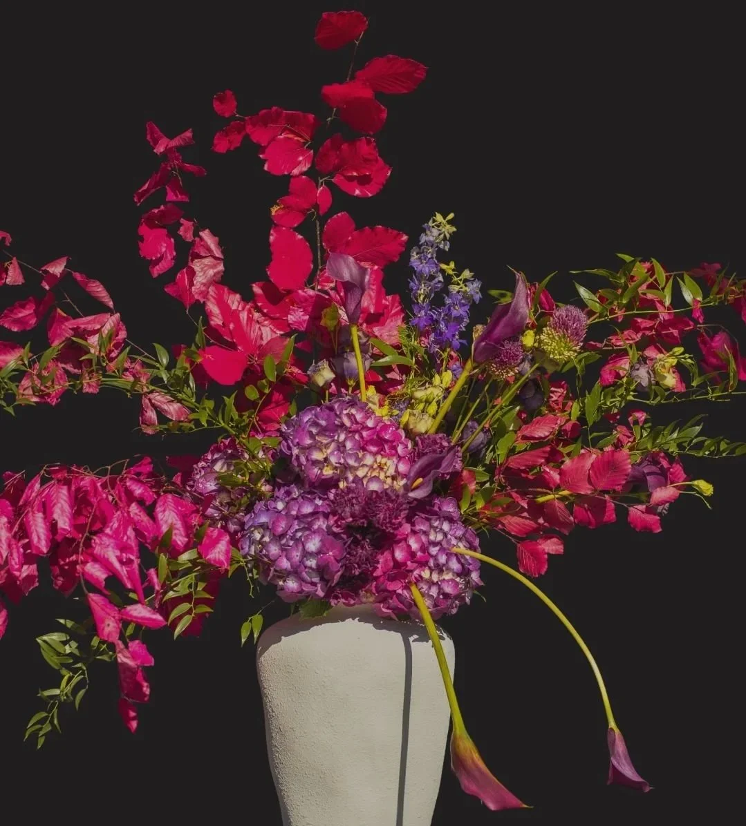 A colorful flower arrangement with pink, purple, and blue flowers in a white vase against a black background.