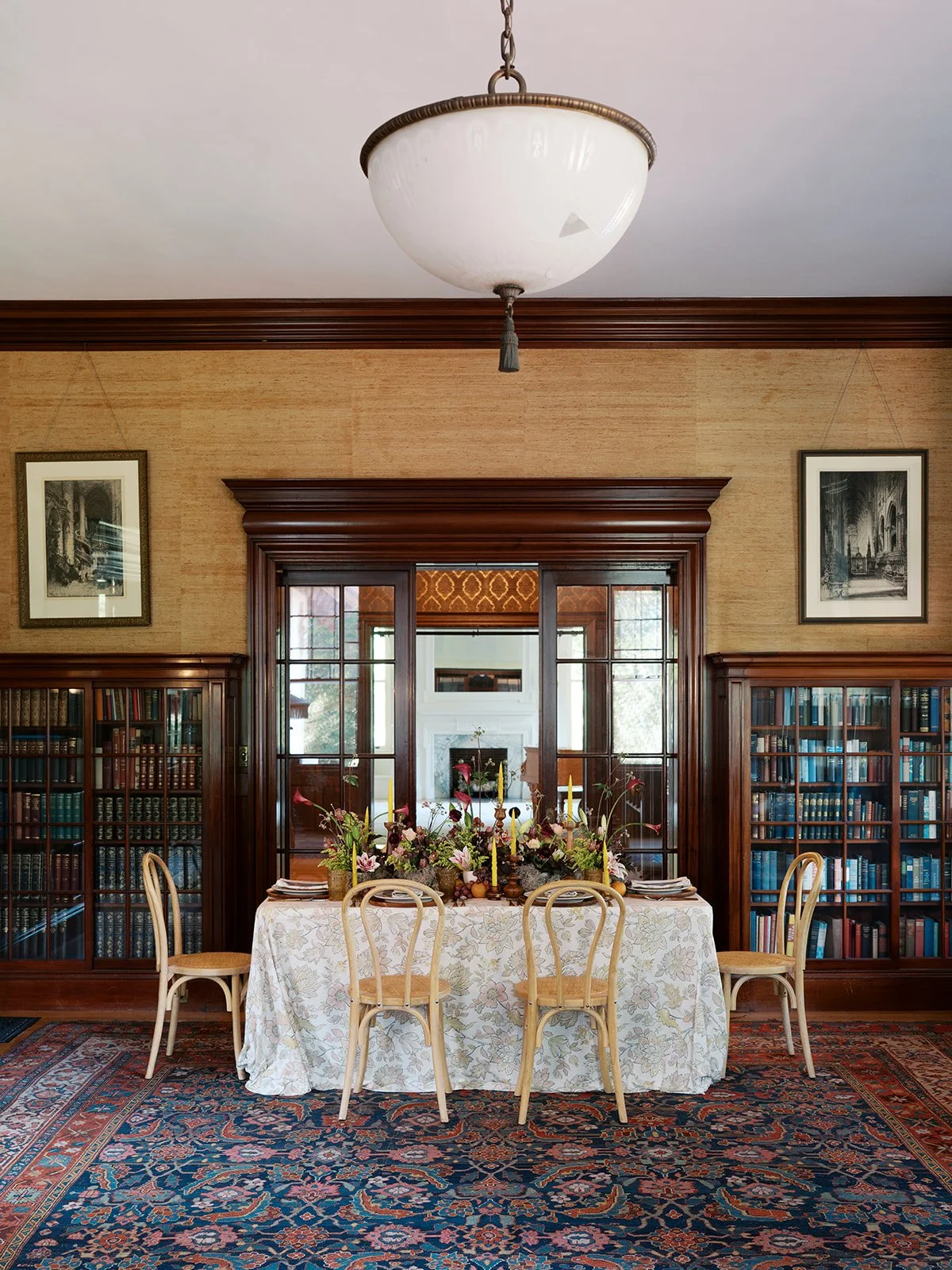 A dining room with a table, six chairs, and floral arrangements, flanked by wooden bookshelves, with a large chandelier overhead, framed pictures on the walls, and a view into an adjacent room with a fireplace.