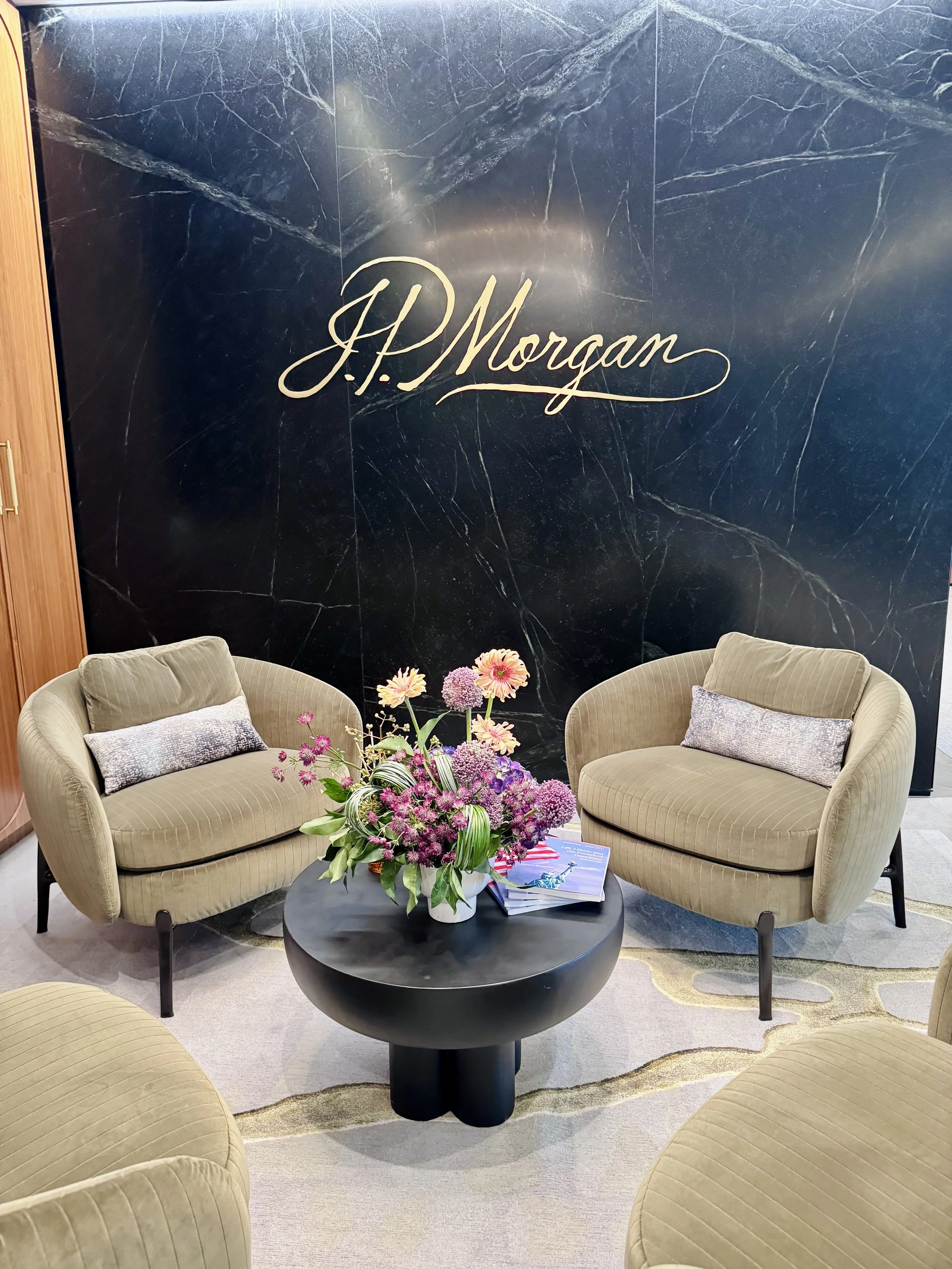 A seating area with six beige armchairs surrounding a black round coffee table topped with a pink and purple flower arrangement and a stack of magazines, set against a black marble wall with gold script lettering that reads 'J.P. Morgan'.