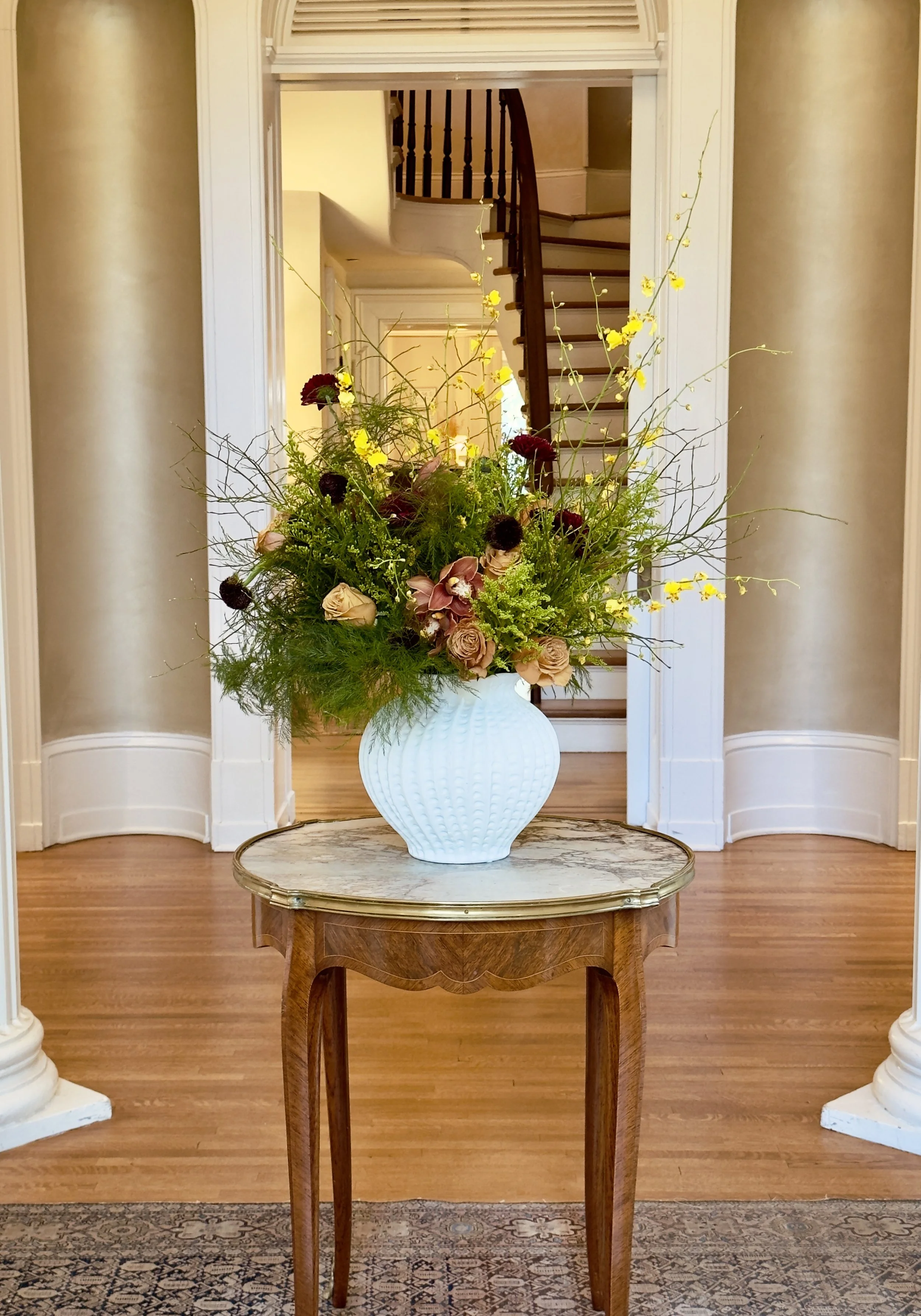 A large floral arrangement in a white textured vase on a wooden table in front of a hallway with a staircase and beige walls.