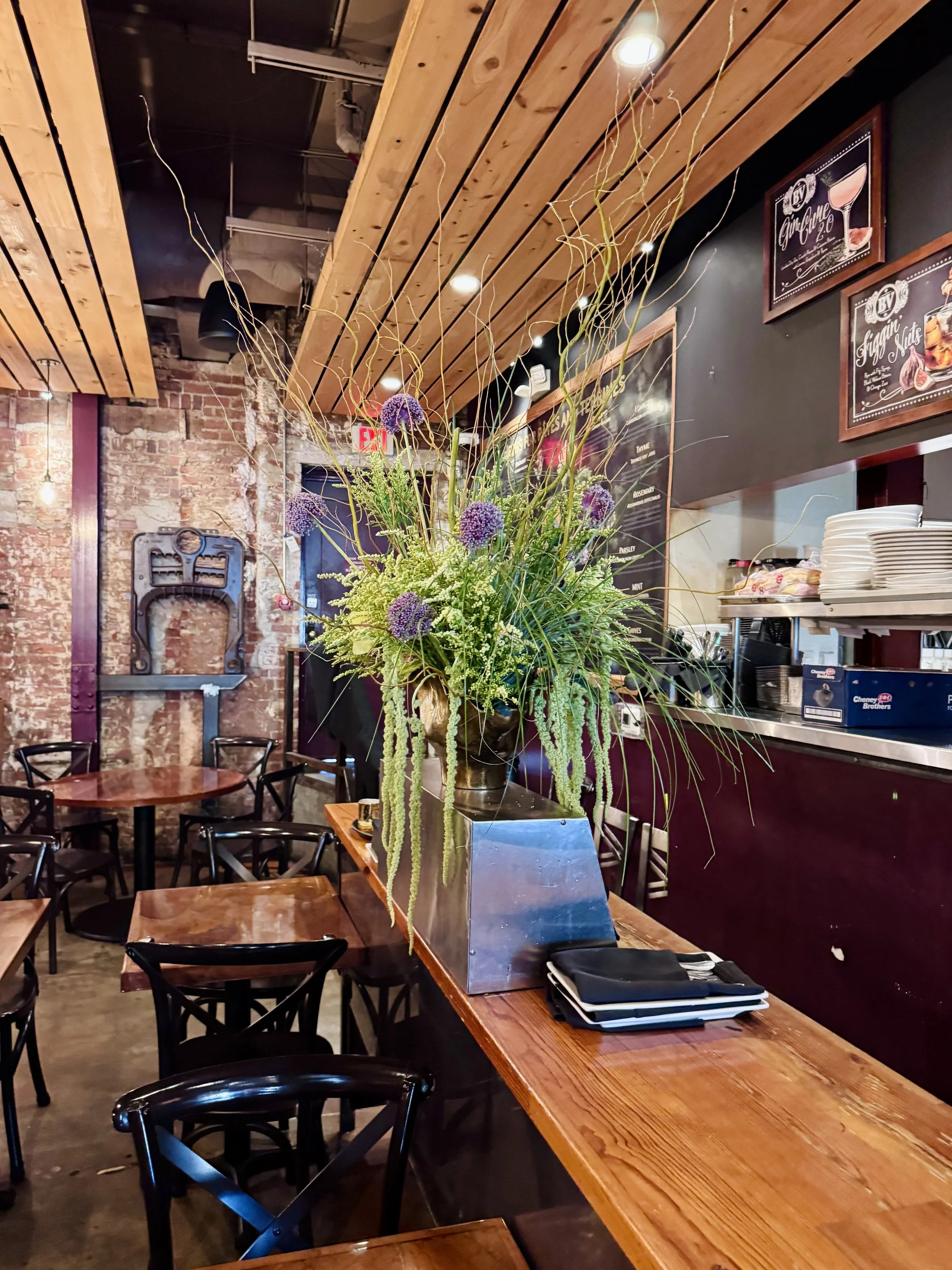 Interior of a restaurant with a large floral arrangement on a wooden table, featuring purple flowers and green hanging plants. Surrounding tables and chairs are visible, with exposed brick wall and wooden ceiling panels.