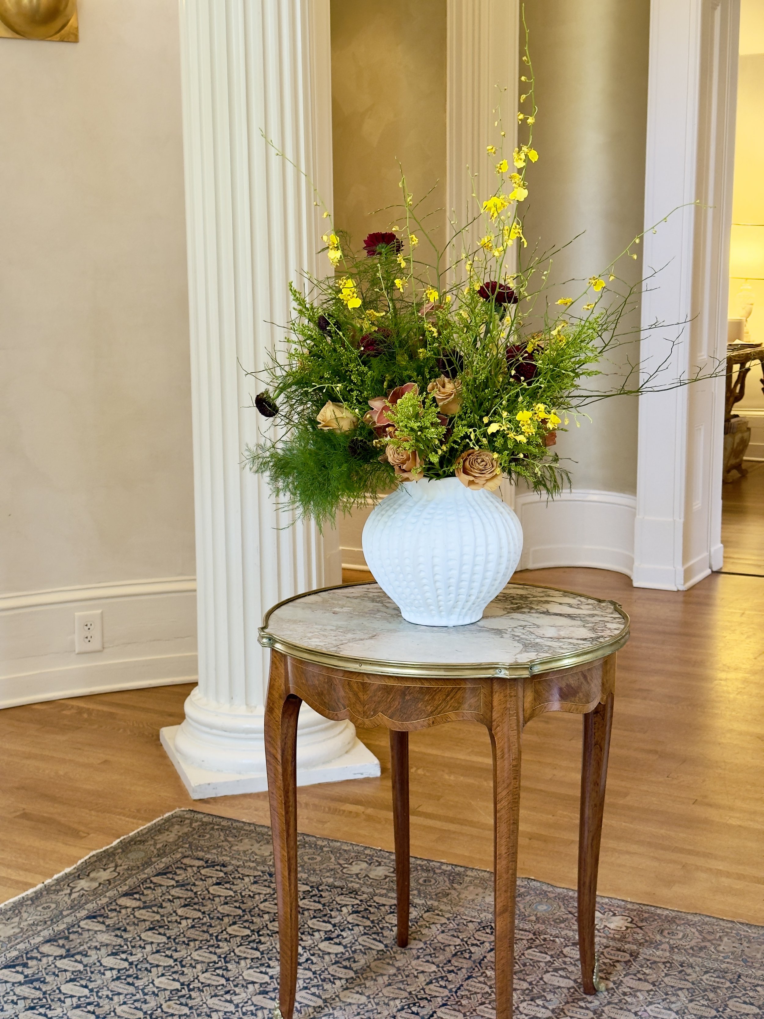 A floral arrangement with beige roses, dark red flowers, and yellow flowers in a white vase with textured patterns, placed on a vintage wooden table with curved legs, in a room with a patterned rug, wooden floor, beige walls, and white decorative col