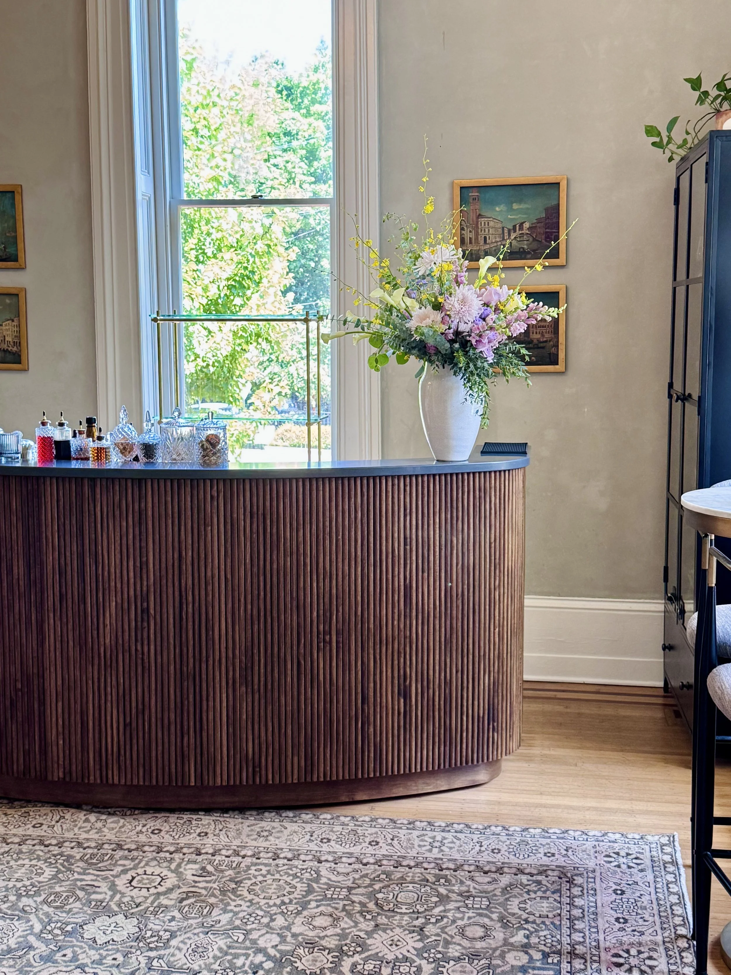 Indoor scene with a wooden curved reception desk, a floral arrangement in a white vase on the desk, a window with lush green trees outside, framed artwork on the wall, a shelf with bottles and jars behind the desk, and a patterned area rug on the hardwood floor.