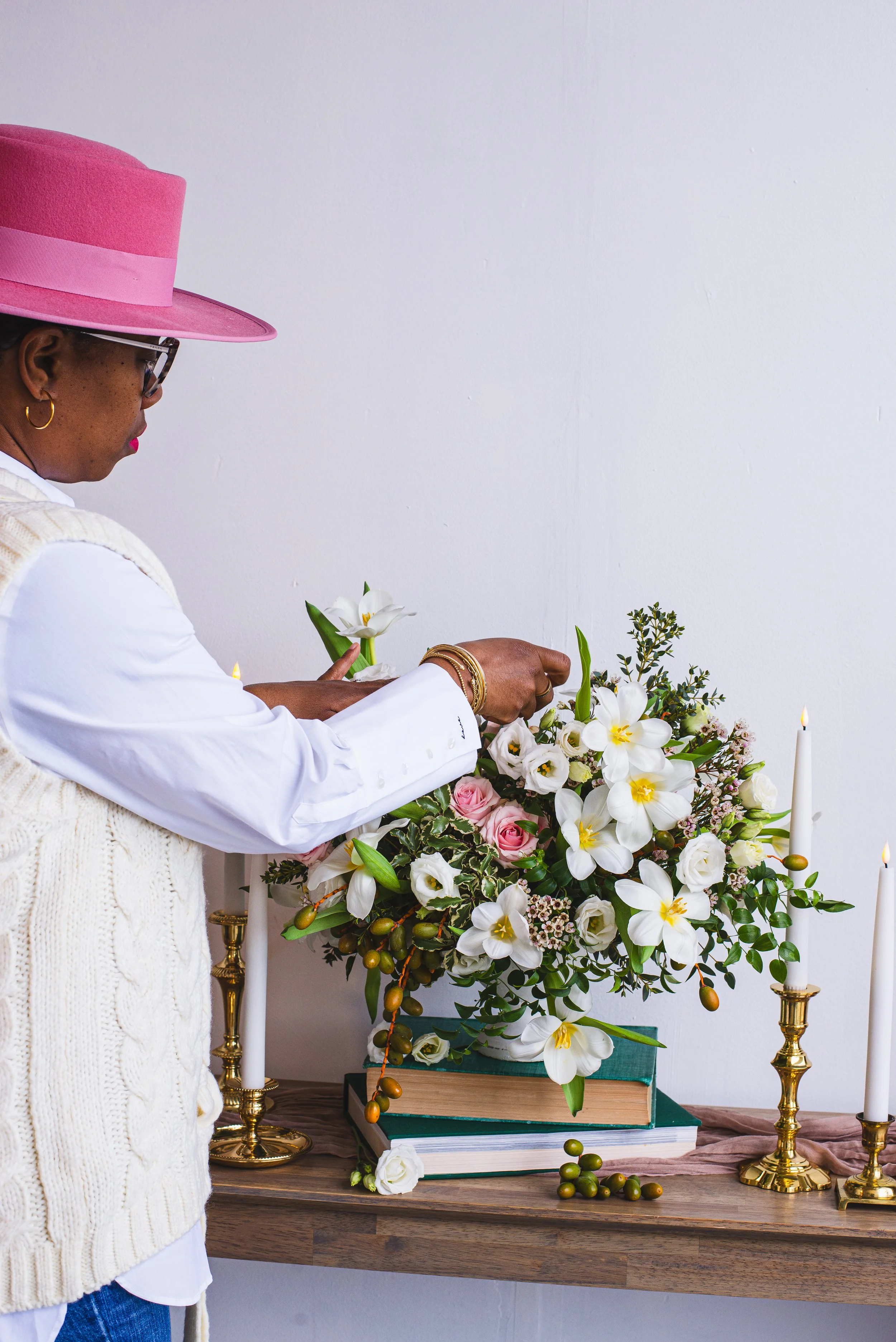 Woman arranging a floral arrangement with white and pink flowers on a table with candles and books.