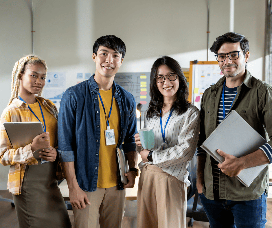 A diverse group of four young professionals standing together in an office, smiling at the camera, holding folders, tablets, and a coffee mug.