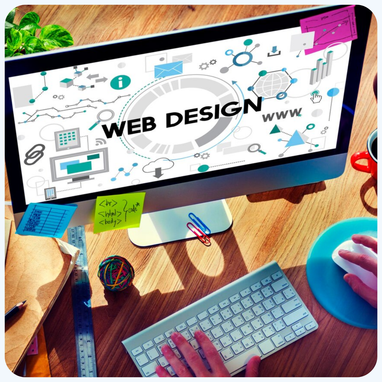 Computer screen displaying a web design concept with various icons and graphics. The workspace includes a keyboard, mouse, ruler, sticky notes, and office supplies on a wooden desk.