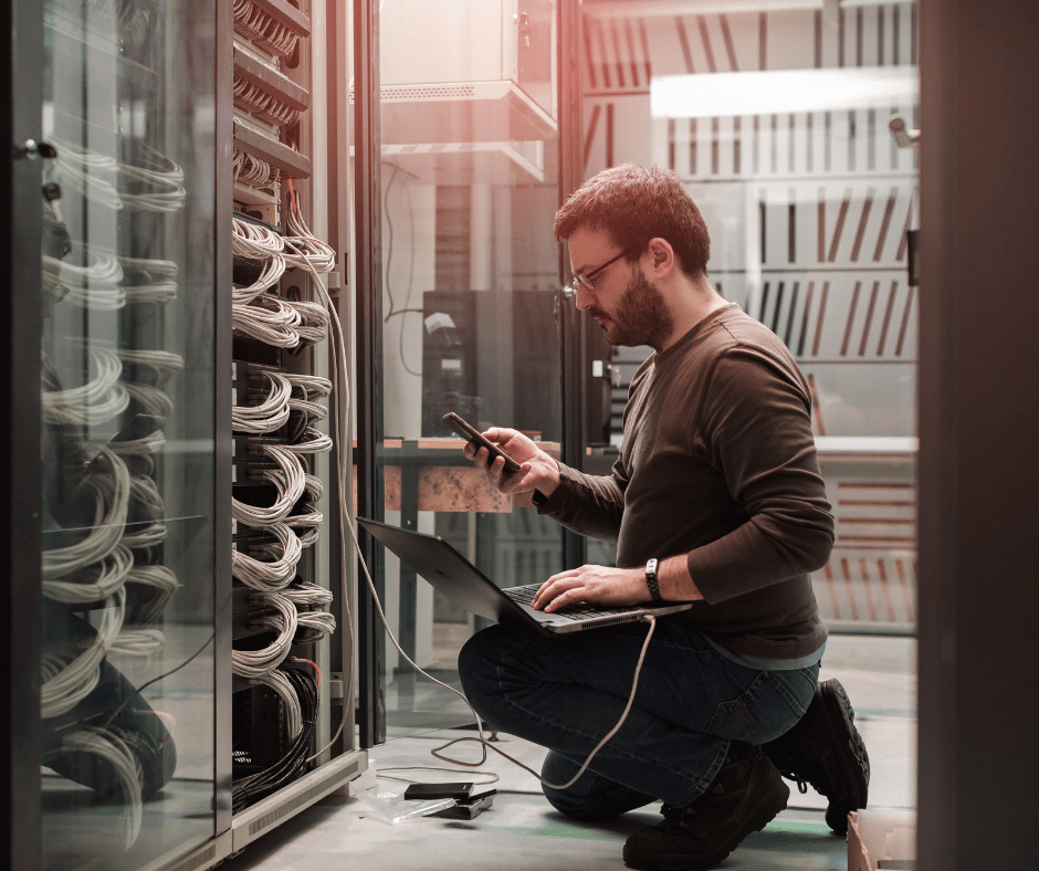 A man in a server room working with networking equipment, using a laptop and smartphone.