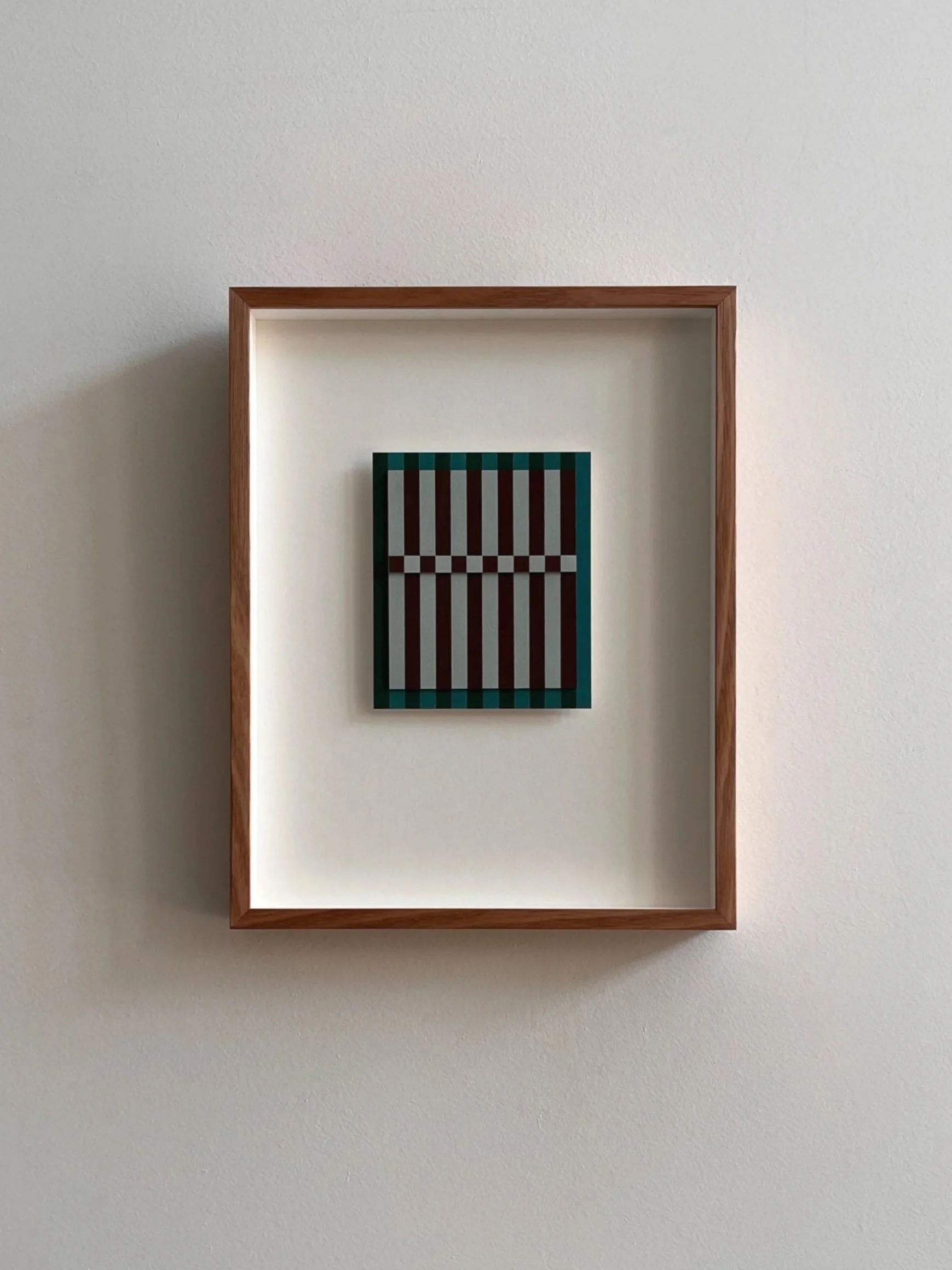 Minimalist art piece in a wooden shadow box with black, white, and teal geometric striped pattern on a white wall.