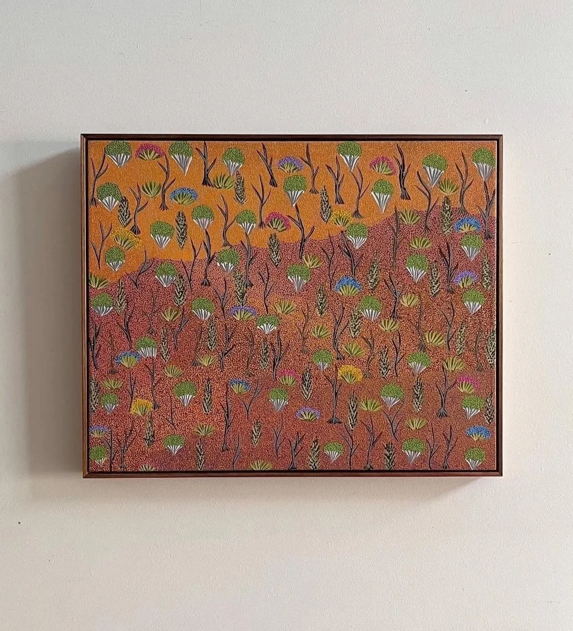 Colorful abstract painting of trees in various green, yellow, purple, and blue shades, set against an orange and red background, framed and hanging on a white wall.