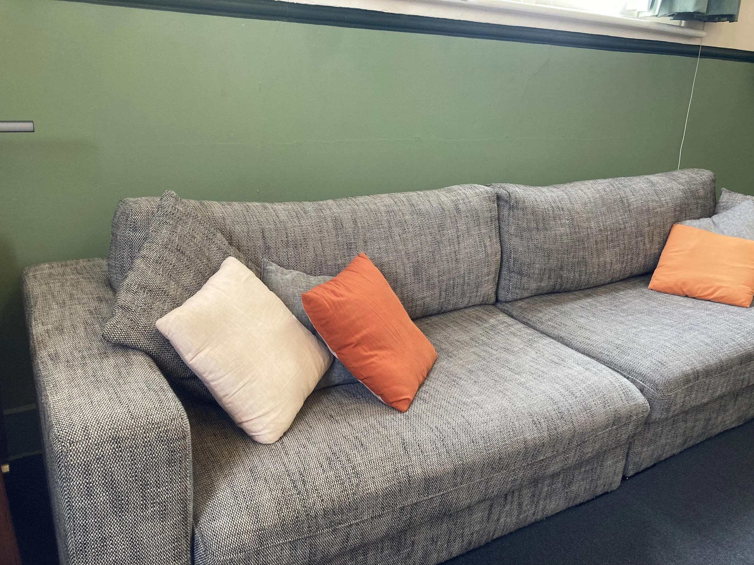 The therapy couch is a gray fabric couch with four cushions, three in orange and one in off-white, placed against a green wall with a window above.