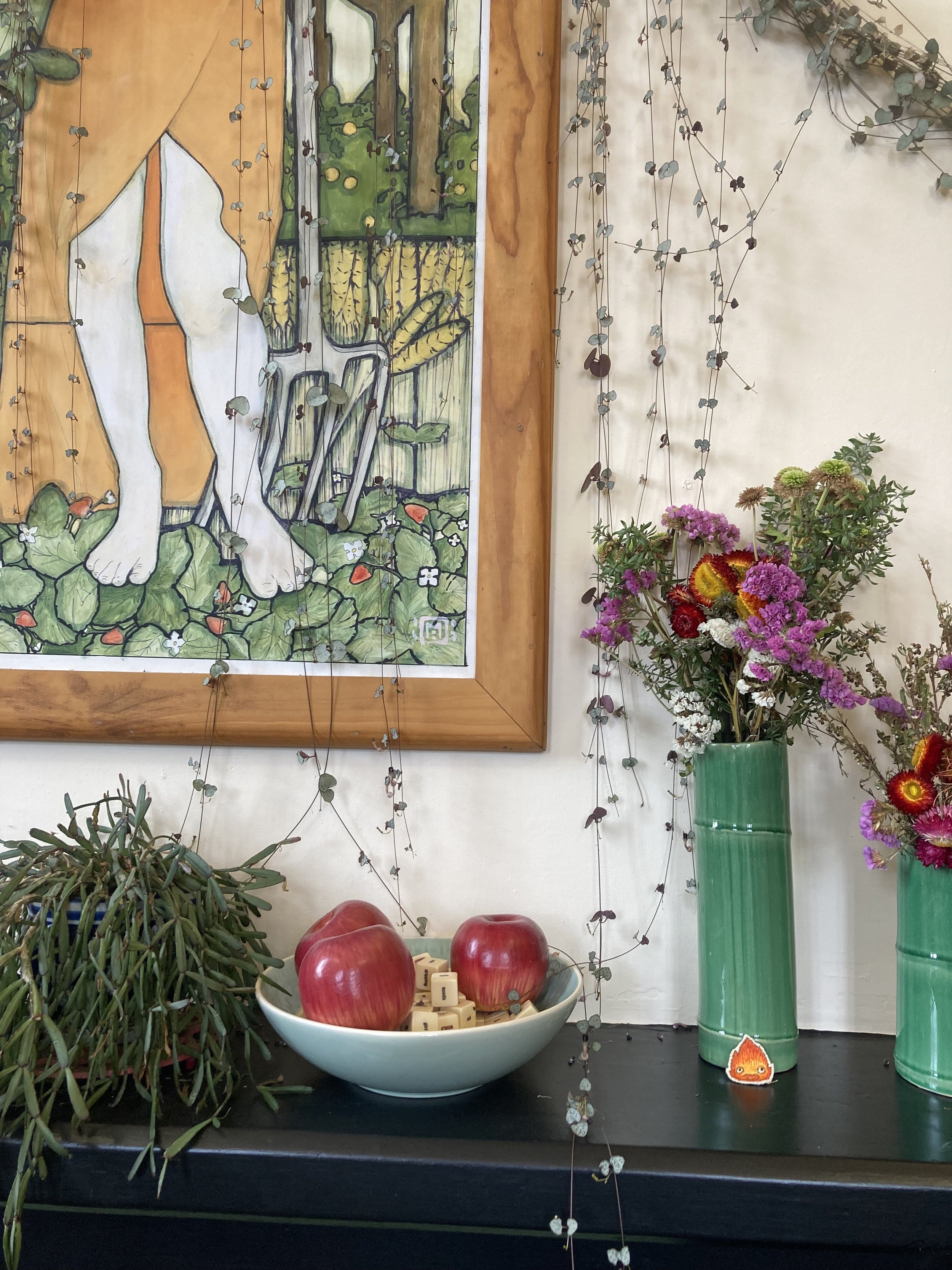 Decorative display with potted succulents, a bowl with three red apples and small wooden blocks, and two green vases filled with purple and pink flowers, positioned on a black surface against a wall decorated with hanging dried vines and a framed artwork of cat paws and garden scene.