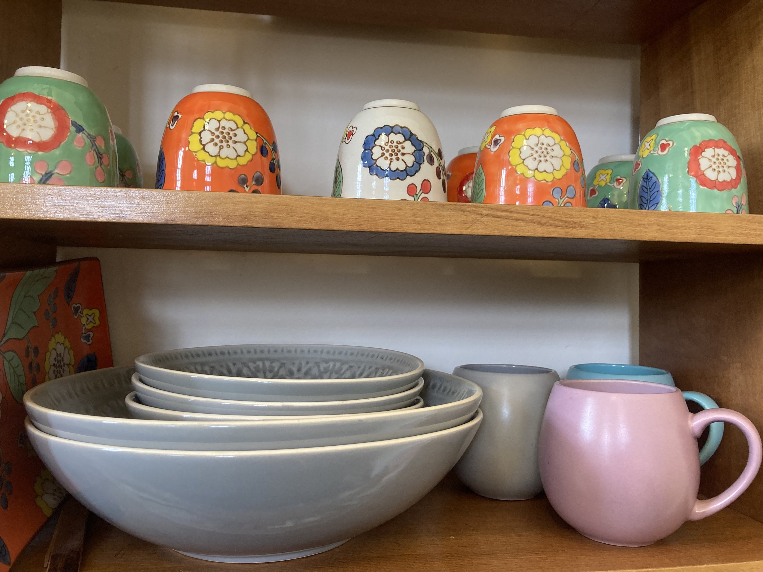 Kitchen shelf with colorful ceramic cups and plates, including floral patterned cups on the top shelf and plain mugs and bowls on the bottom shelf.