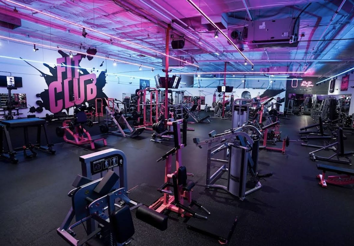 Interior of a gym with pink and blue neon lighting, featuring various workout equipment and a large mural that says 'Fit Club' on the wall.