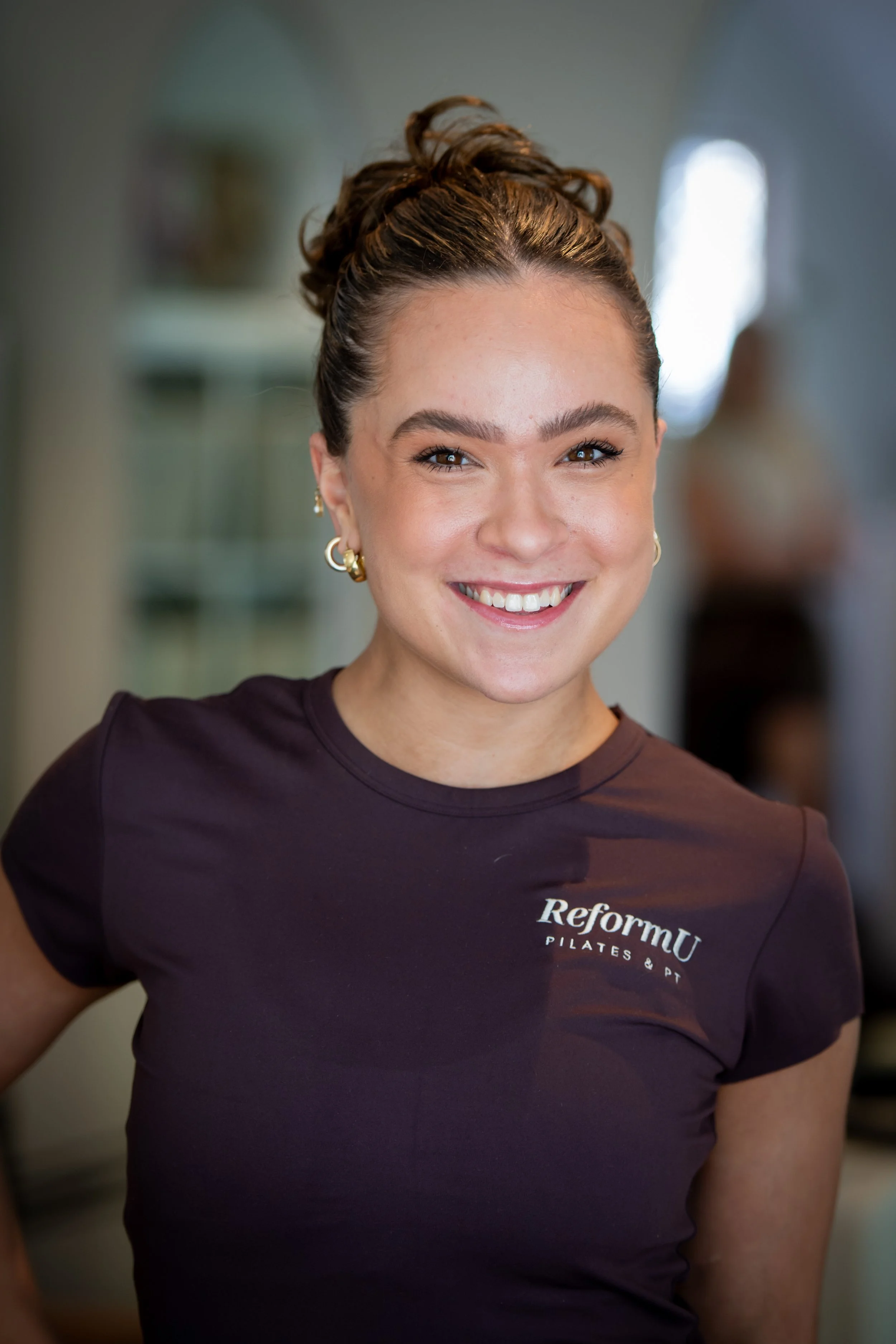 A young woman with brown hair styled with black bows, smiling and wearing a dark purple ReFornu Pilates & PT t-shirt, standing indoors with a blurred background.