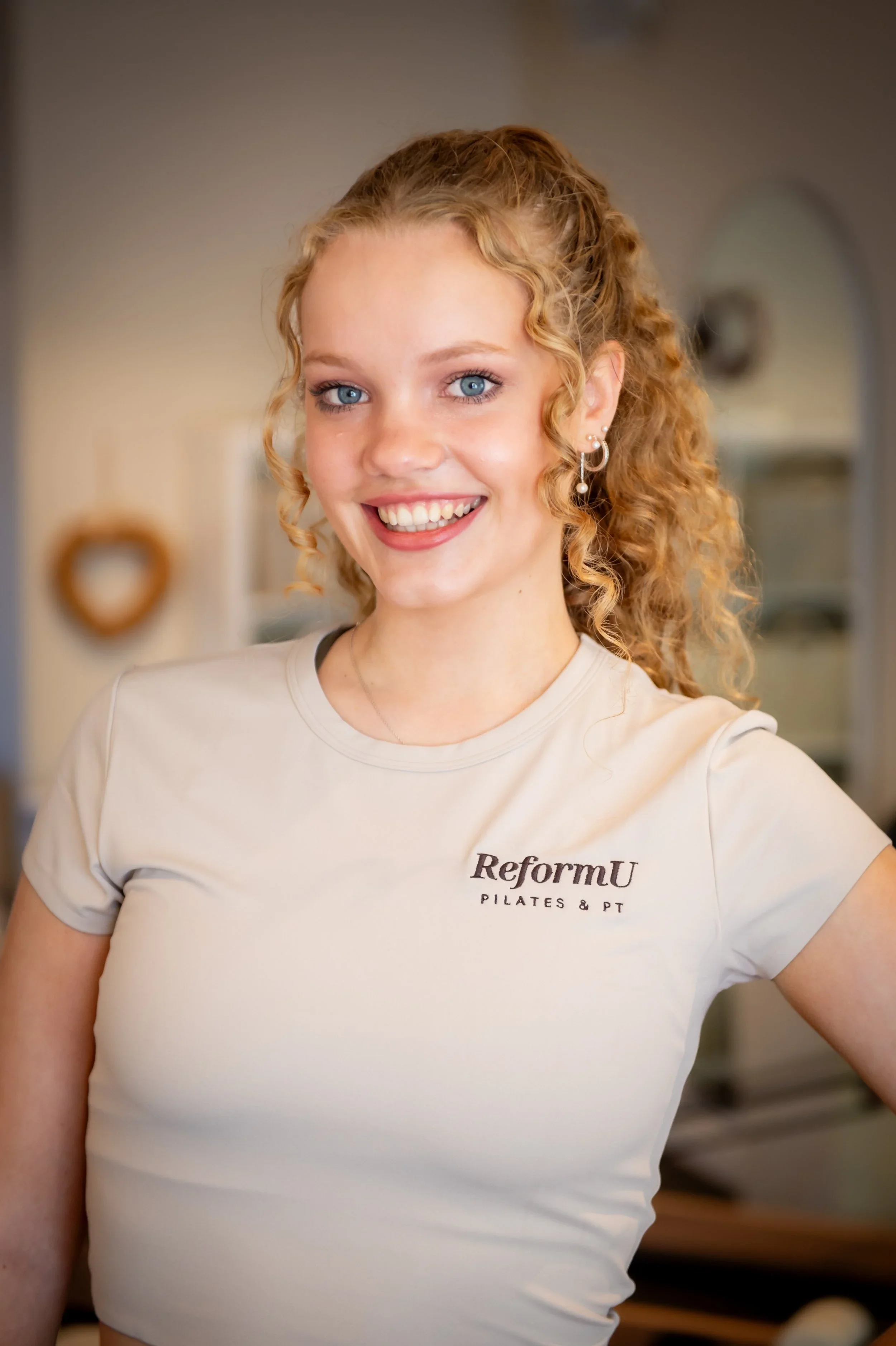 A young woman with curly blonde hair and blue eyes, smiling, wearing a beige ReforU Pilates & PT T-shirt, standing inside a home or studio with blurred background.