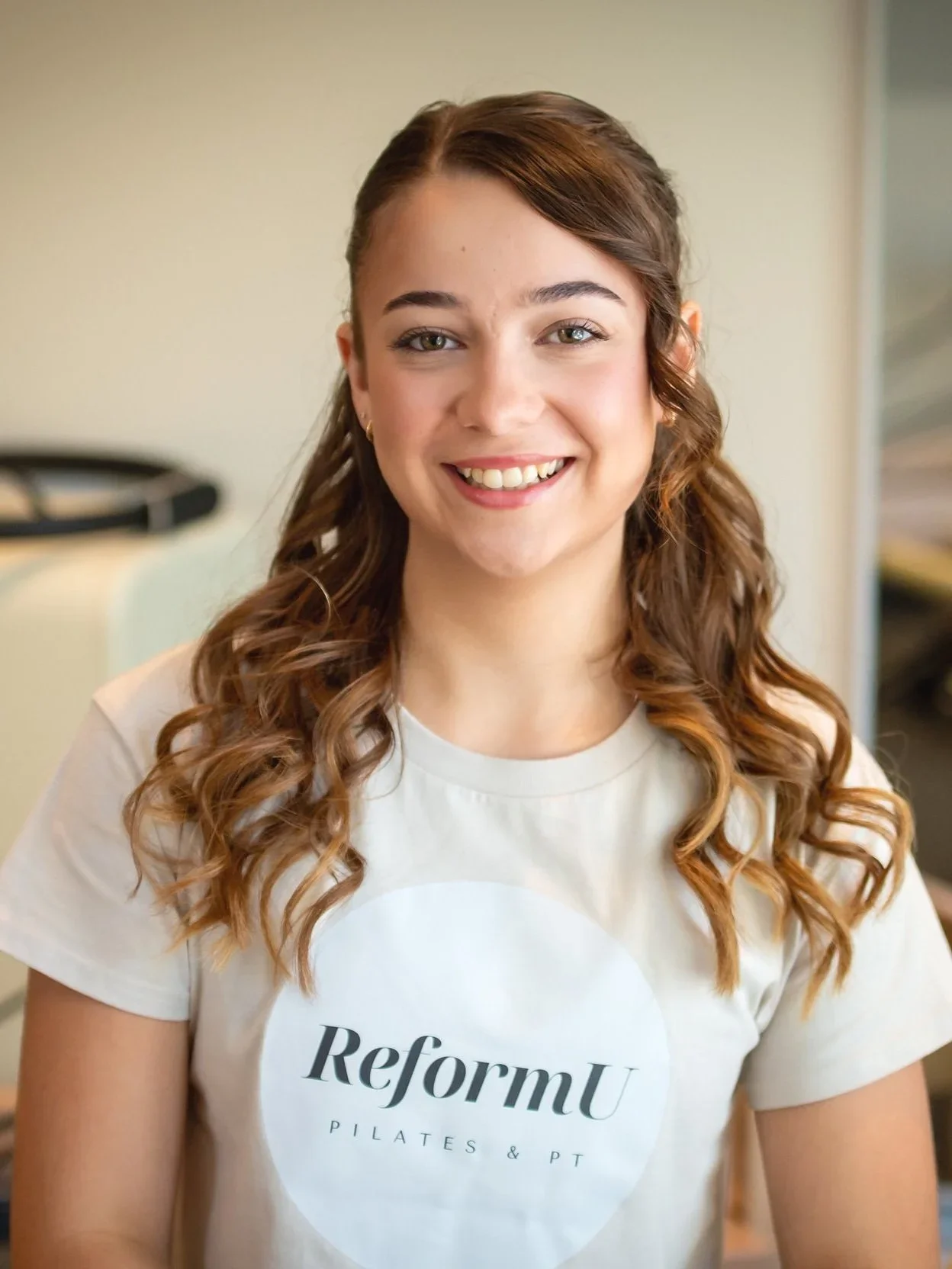 Young woman with long, curly brown hair smiling, wearing a white ReformU Pilates & PT T-shirt.