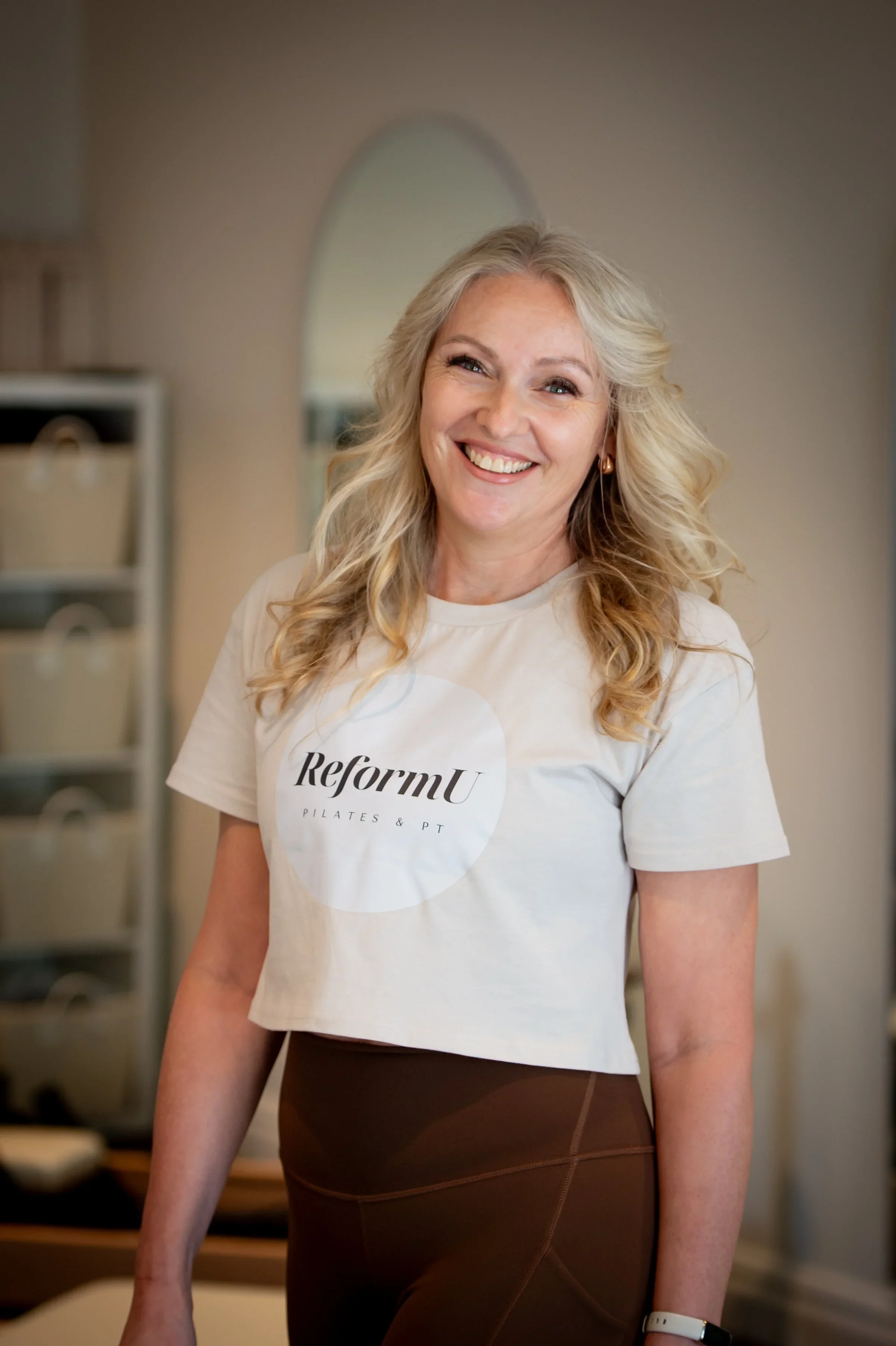 A woman with blonde, wavy hair smiling in a Pilates studio, wearing a white ReforMu t-shirt and brown leggings.
