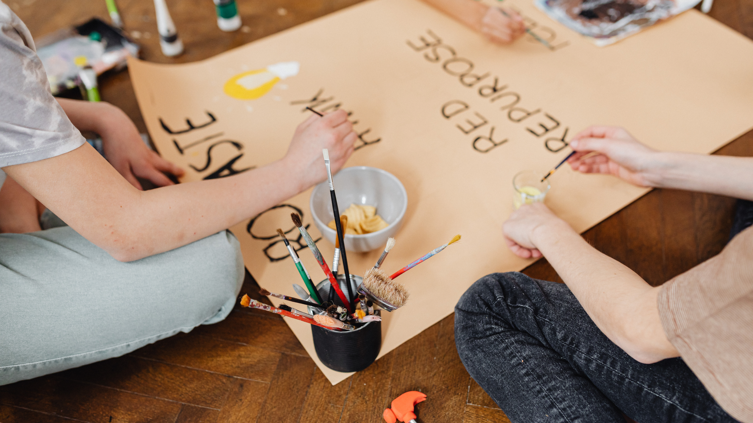 People creating a project poster with black lettering on a large beige poster, surrounded by paintbrushes, paint bottles, a bowl of snacks, and crafting supplies on a wooden floor.