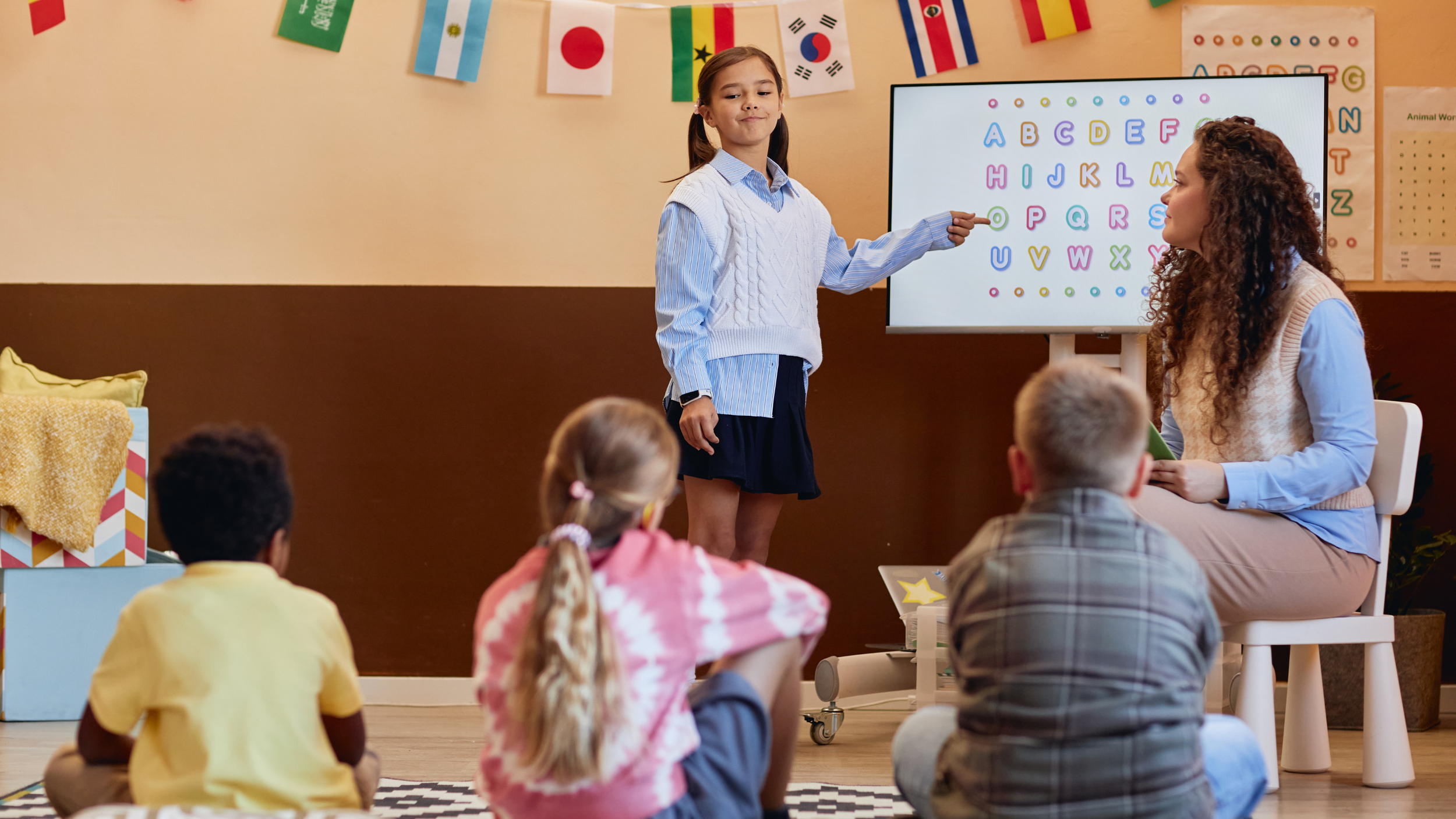 A young girl giving a presentation in a classroom with an alphabet chart on the digital screen behind her. Four children are sitting on the floor watching, and a teacher or instructor is seated nearby.