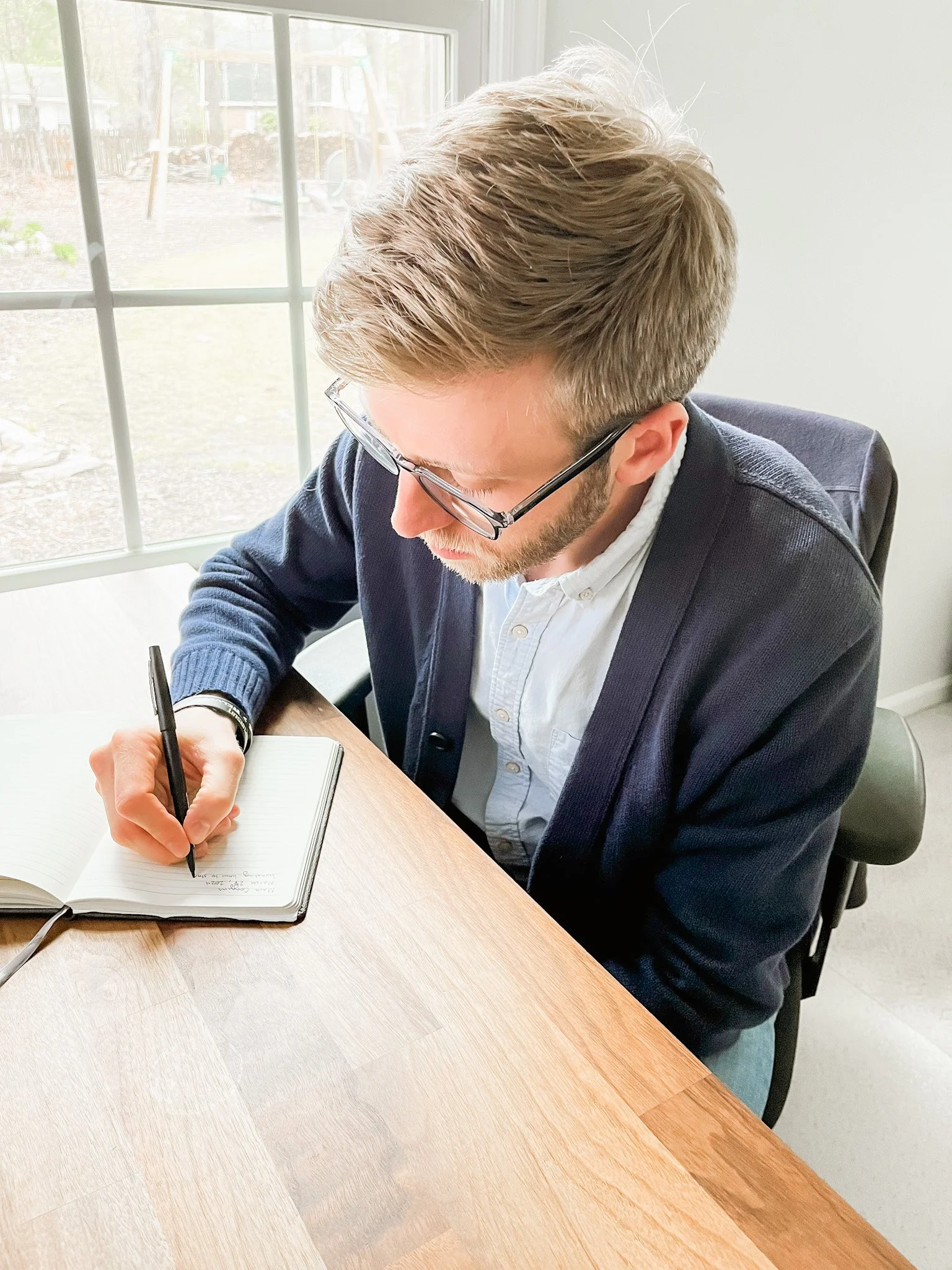 A man with blonde hair and glasses writing in a notebook at a wooden table indoors near a window.