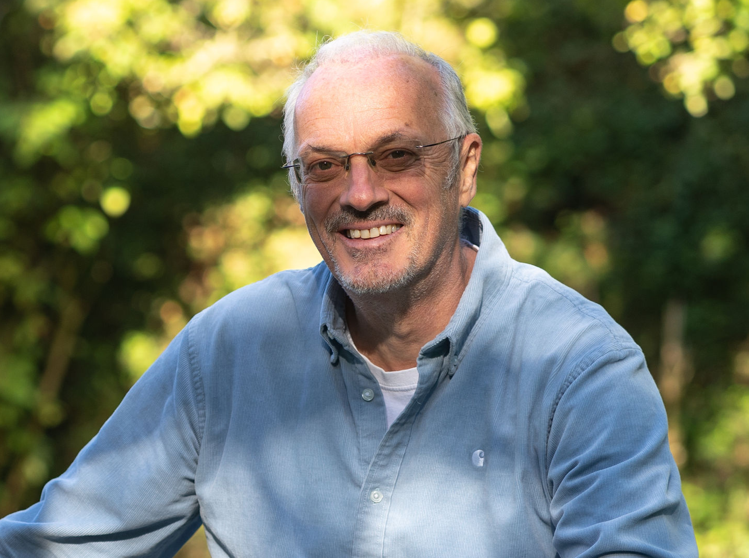 Smiling man wearing glasses and a blue shirt outdoors with greenery in the background.