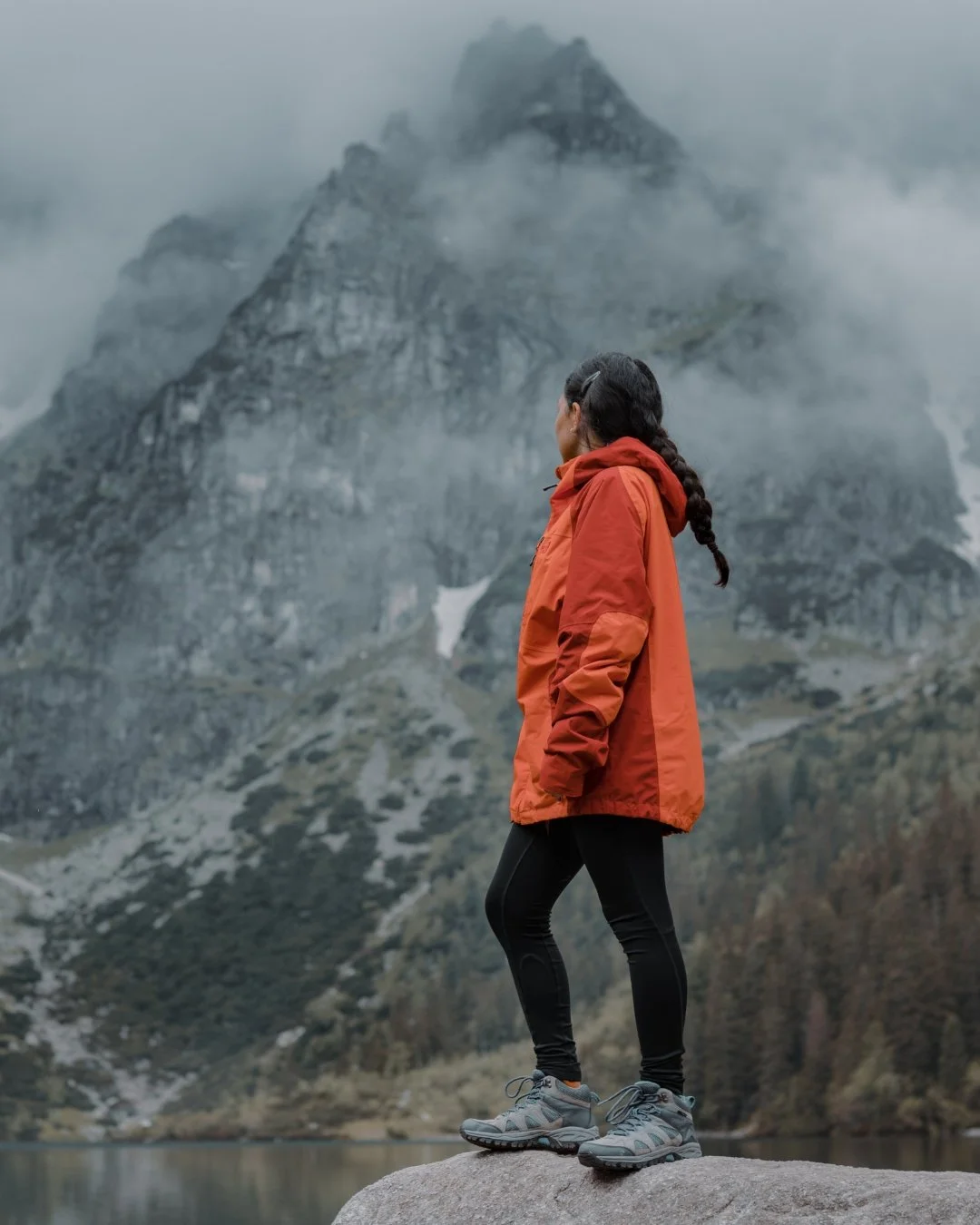 A woman in an orange jacket and black leggings stands on a rock near a lake, looking at fog-covered mountains in the background.