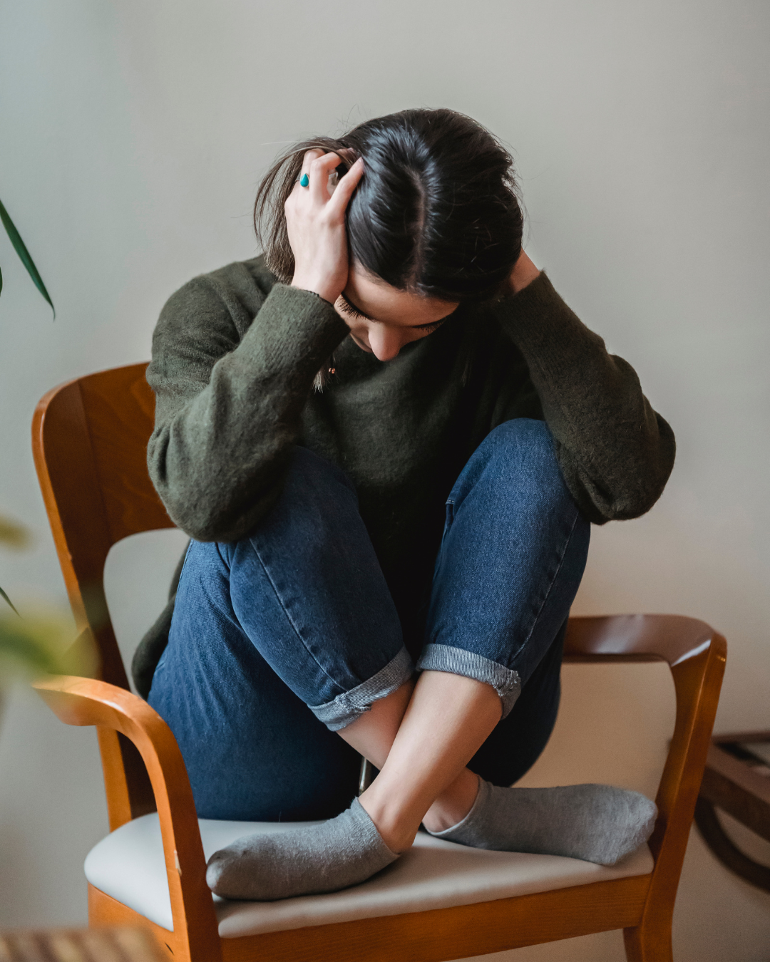 A woman sitting on a chair with her knees drawn up to her chest, holding her head and hair with both hands, appearing distressed or overwhelmed.