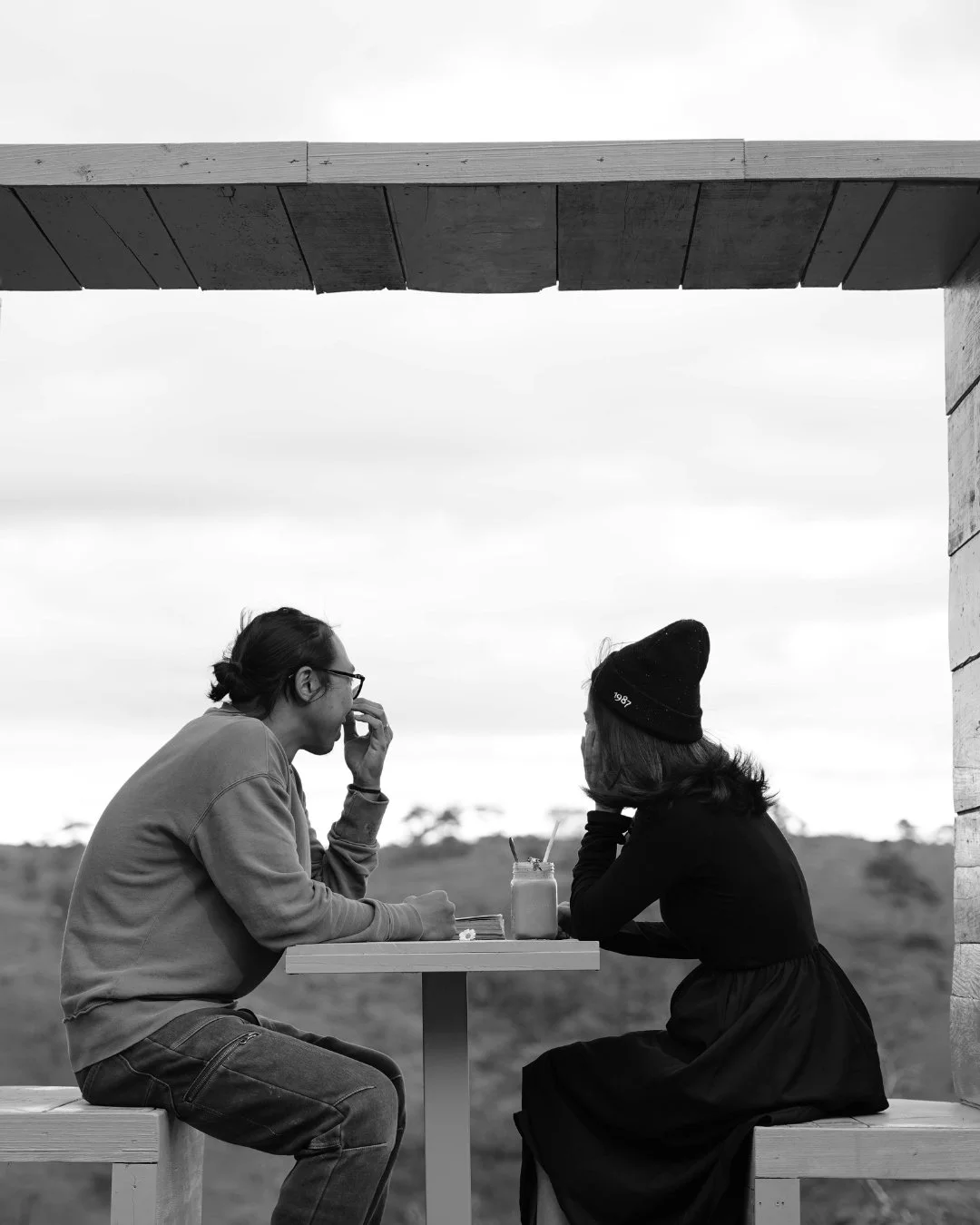 Two people sitting at a table outdoors, engaged in conversation, with drinks and a straw on the table, black and white photo.