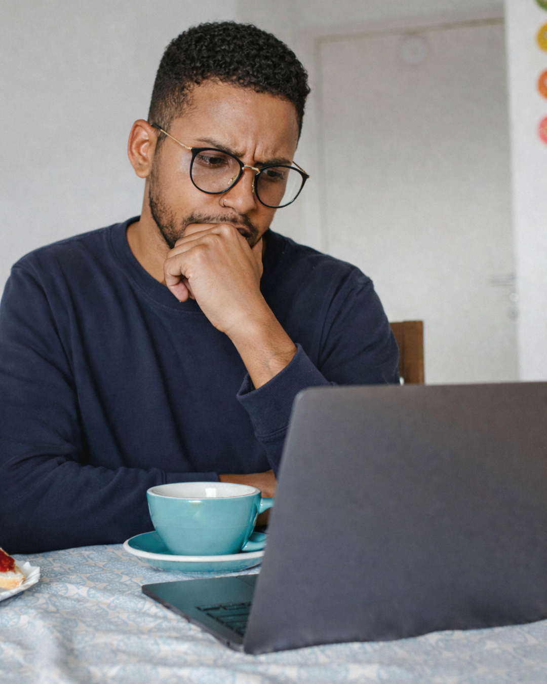 A man wearing glasses and a navy sweater, sitting at a table with a laptop, a blue coffee cup, and a plate with food, looking thoughtful while working or studying.