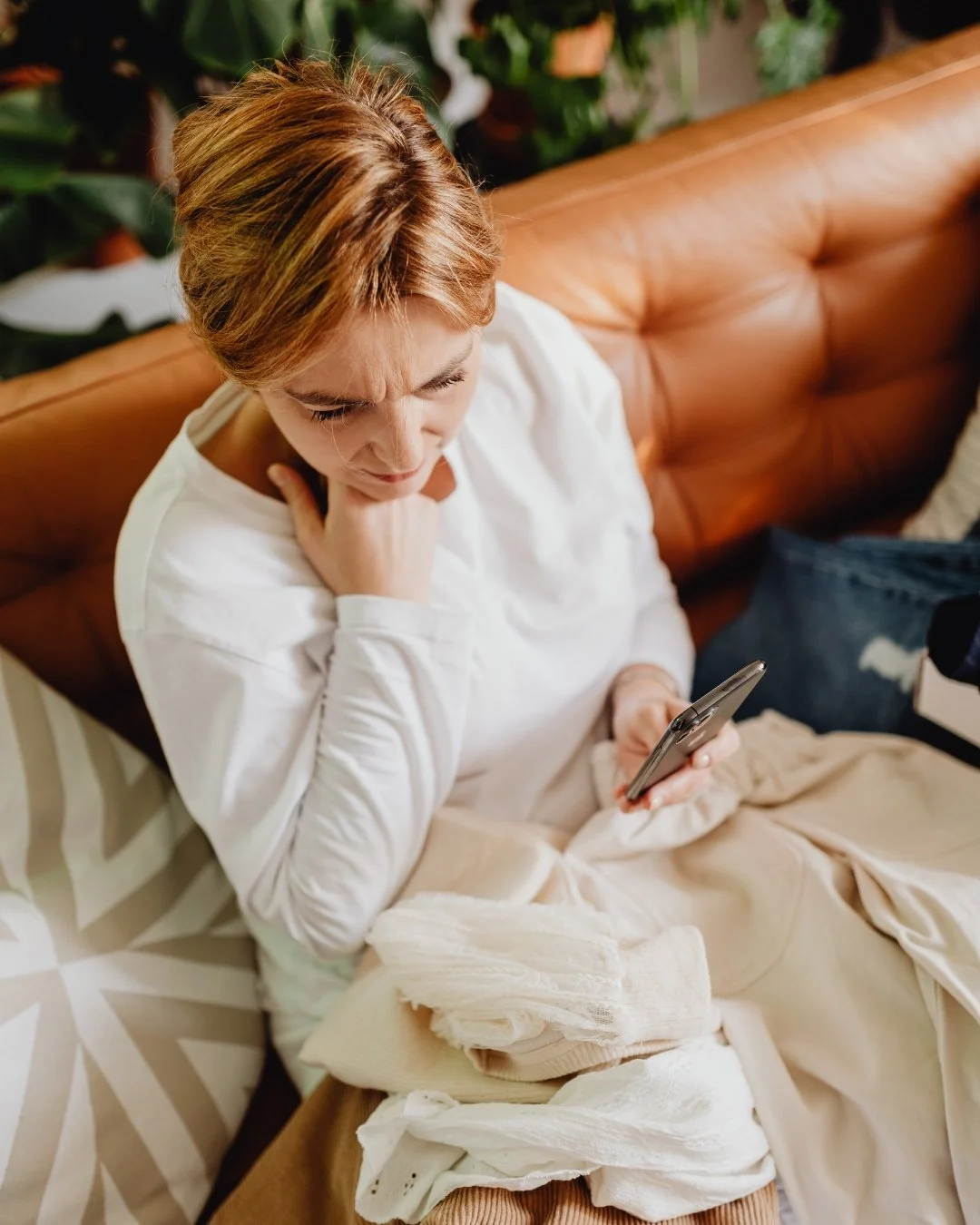 Woman sitting on a sofa looking at her phone, surrounded by clothes.