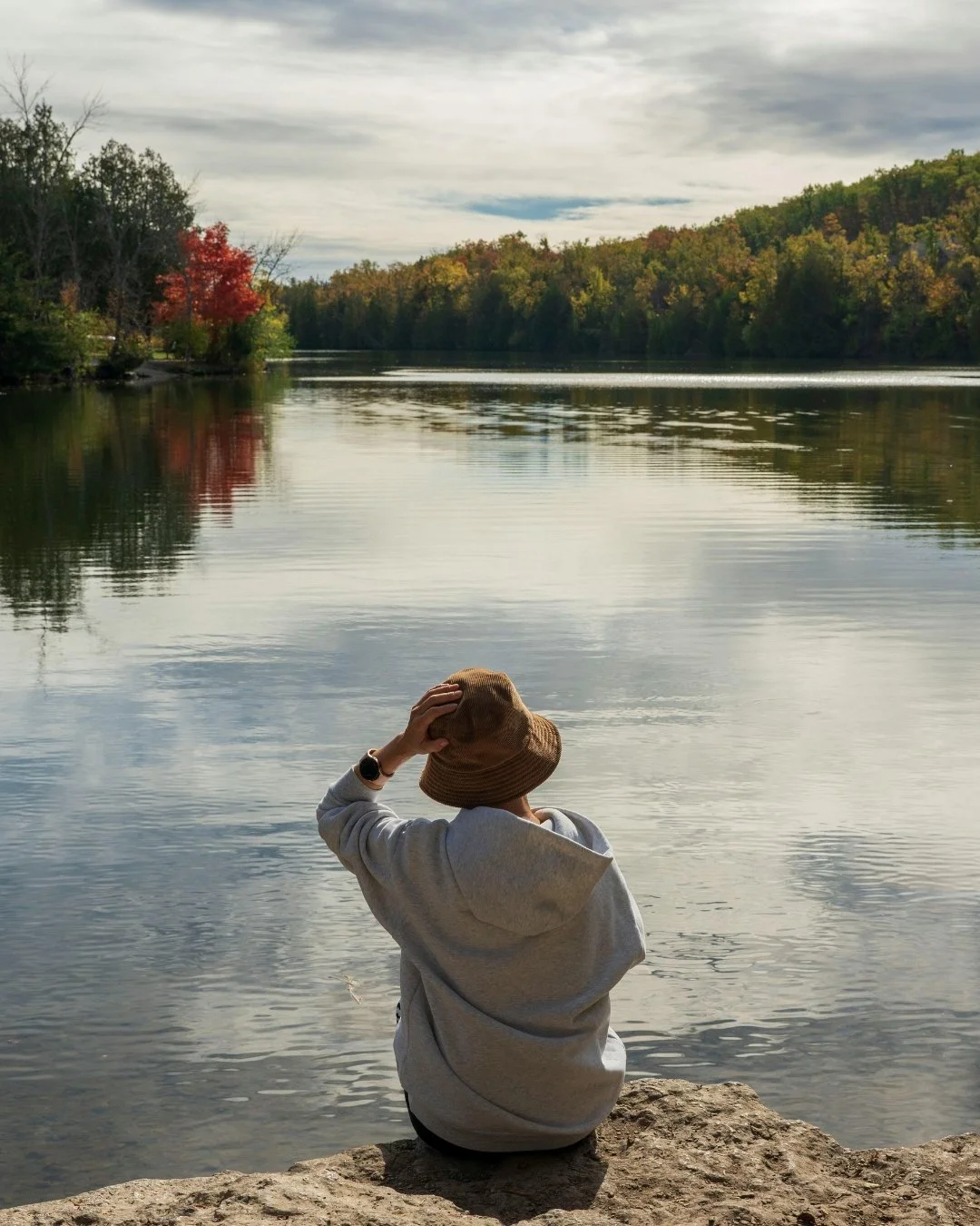 A person wearing a gray hoodie and a brown bucket hat sitting on a rock by a calm river, with trees in autumn colors and a cloudy sky reflected in the water.