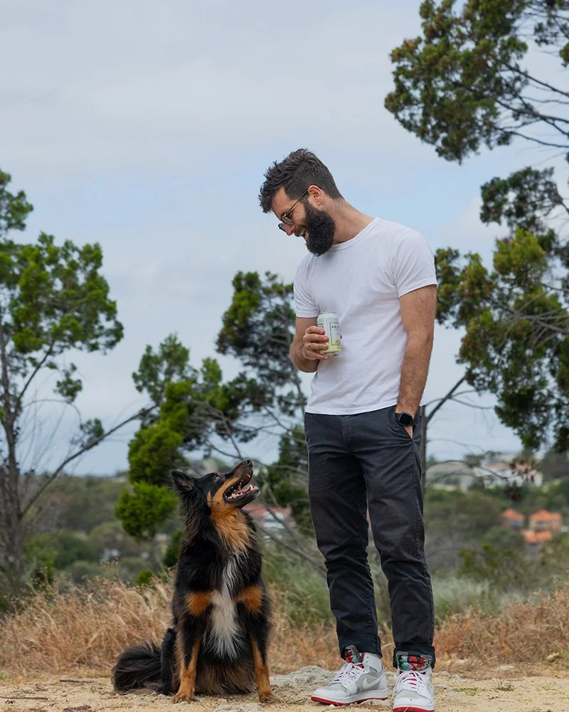 Bearded man enjoying a moment with his dog outdoors, holding a drink – symbolising mindfulness, connection, and living with intention.