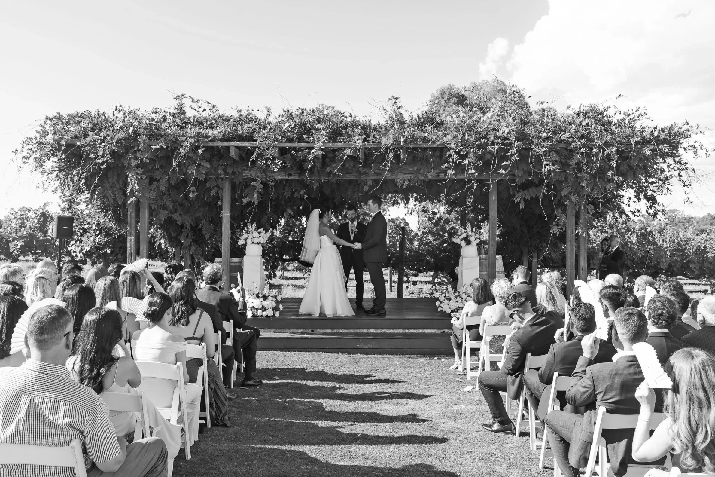 Black and white photo of a wedding ceremony at Sandalford winery with a bride and groom standing on a wooden platform under a leafy arbor, exchanging vows, with guests seated on either side on chairs outdoors and a male celebrant officiating