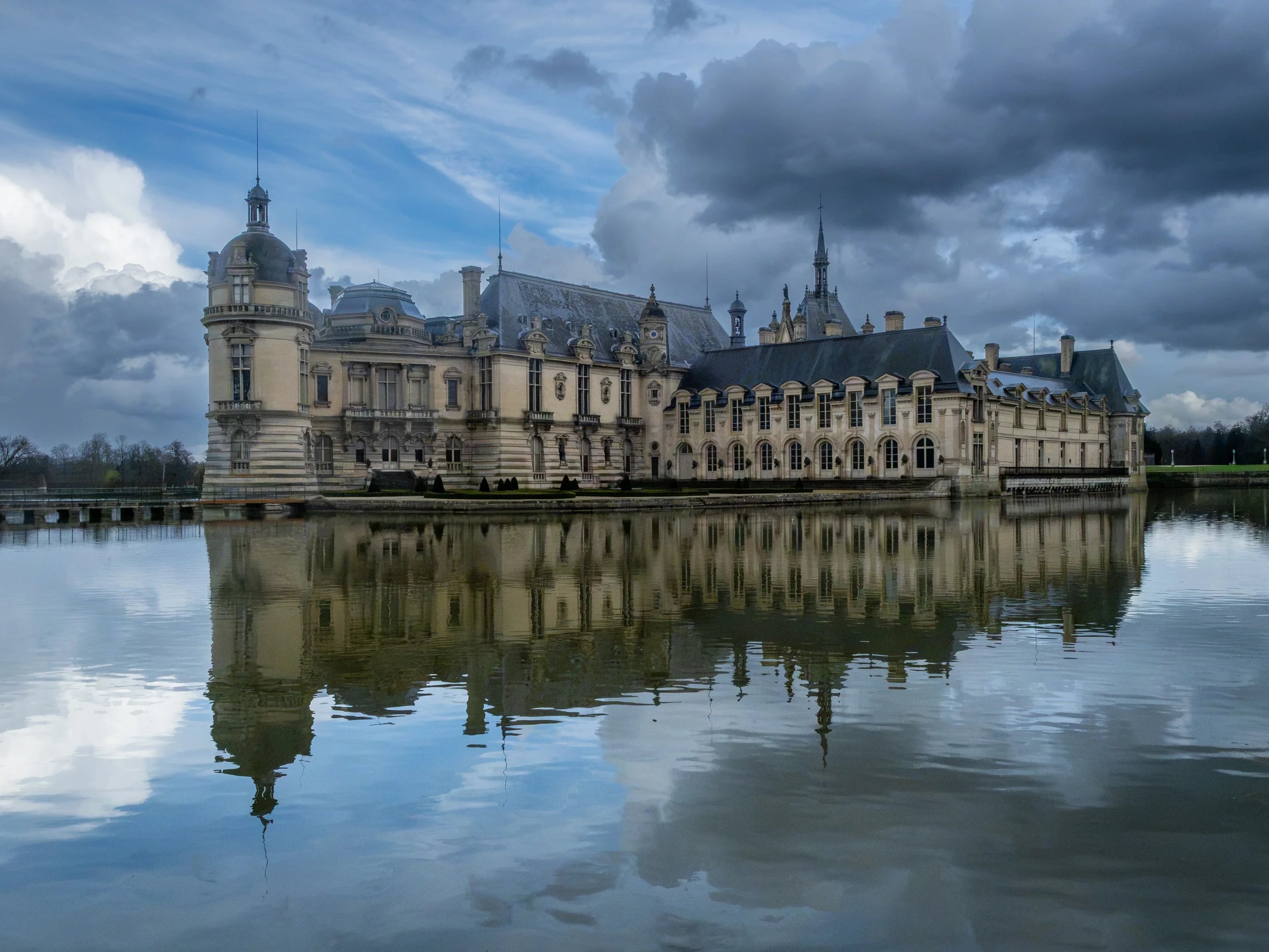 Château Chantilly and its reflection in the surrounding lake, under a cloudy sky.