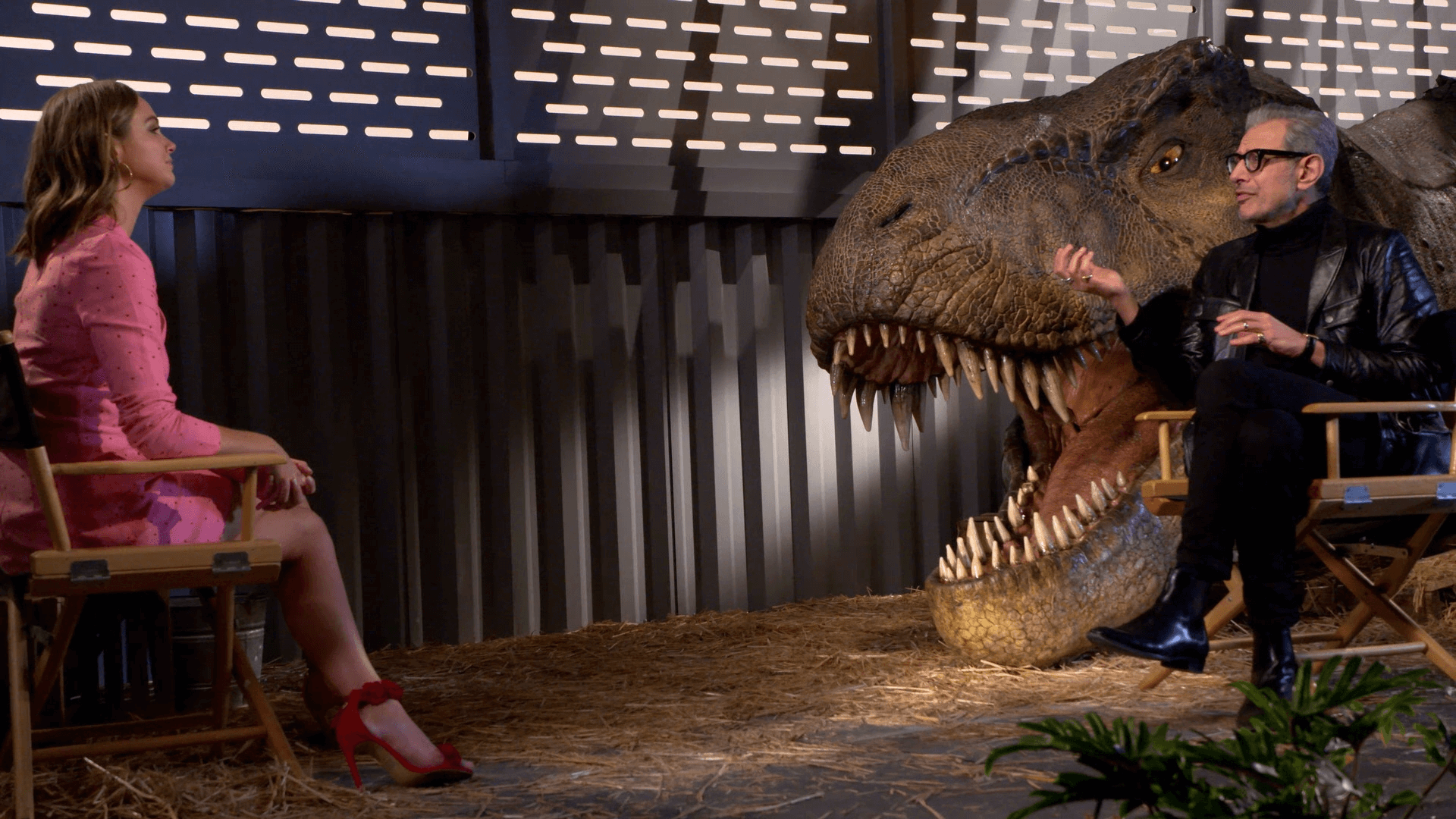 A woman in a pink dress and red high-heeled shoes sitting in a director's chair facing a man with gray hair, glasses, and a black leather jacket. They are seated near a large open-mouthed T-Rex dinosaur model with sharp teeth, inside a room with industrial-style metal walls.