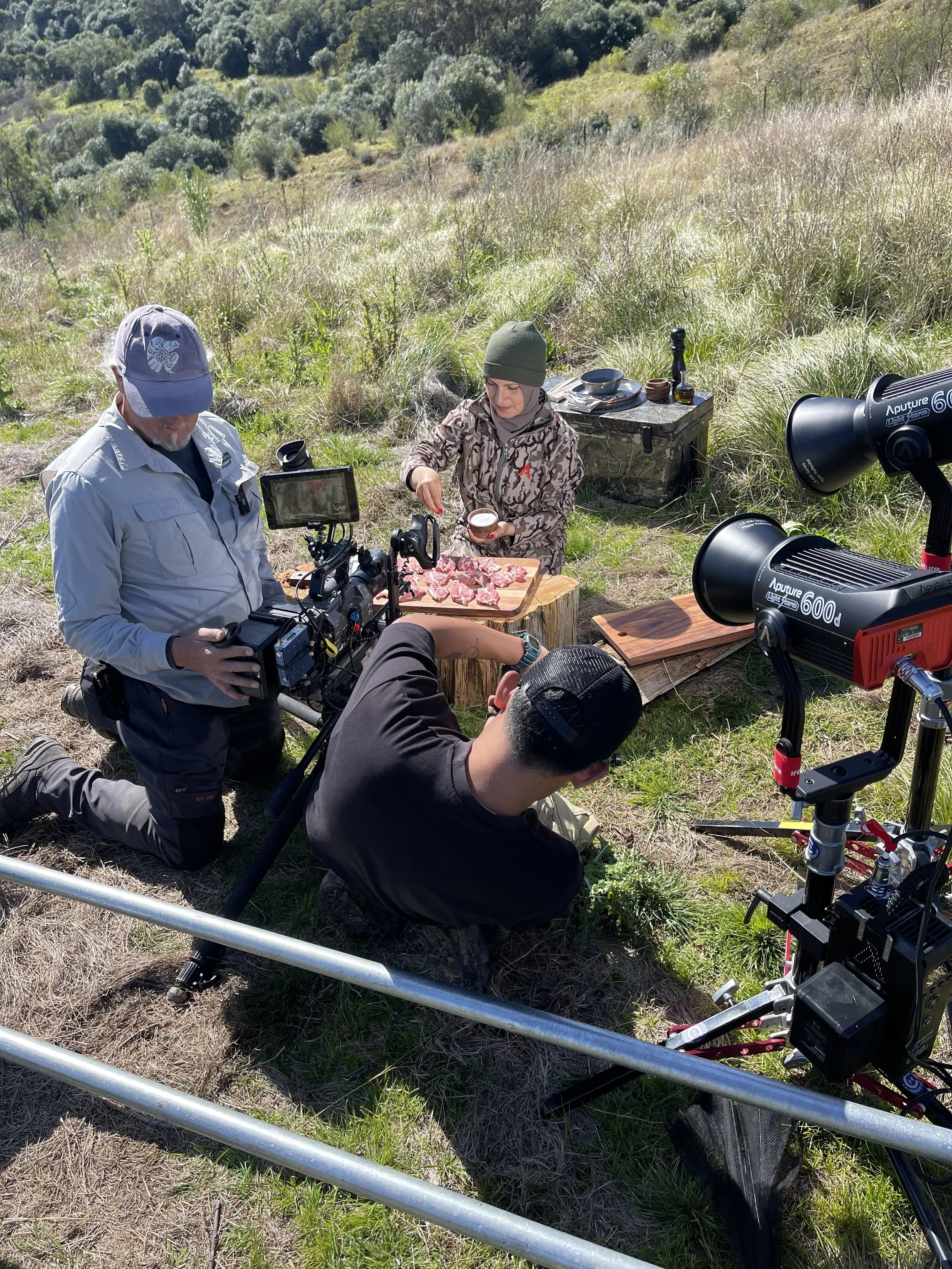 People filming outdoors in a grassy, hilly area with trees in the background. One person is kneeling on the ground, another is standing, preparing meat on a chopping board. Film equipment, including cameras and lights, is set up around them.