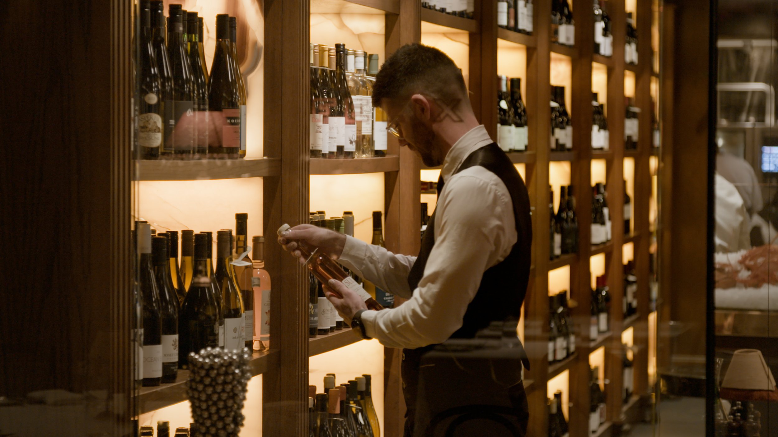 A man with tattoos and glasses examining a wine bottle in a wine cellar or wine shop, surrounded by shelves filled with wine bottles.