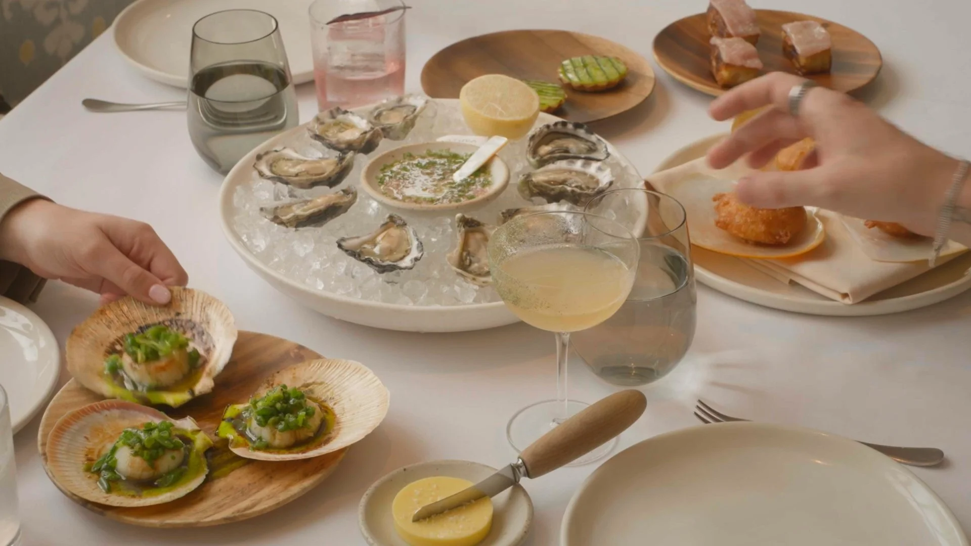 A dining table with oysters on a bed of ice, some garnished with green onions, various small plates with food, a glass of white wine, and a person's hand reaching for a fried food item.