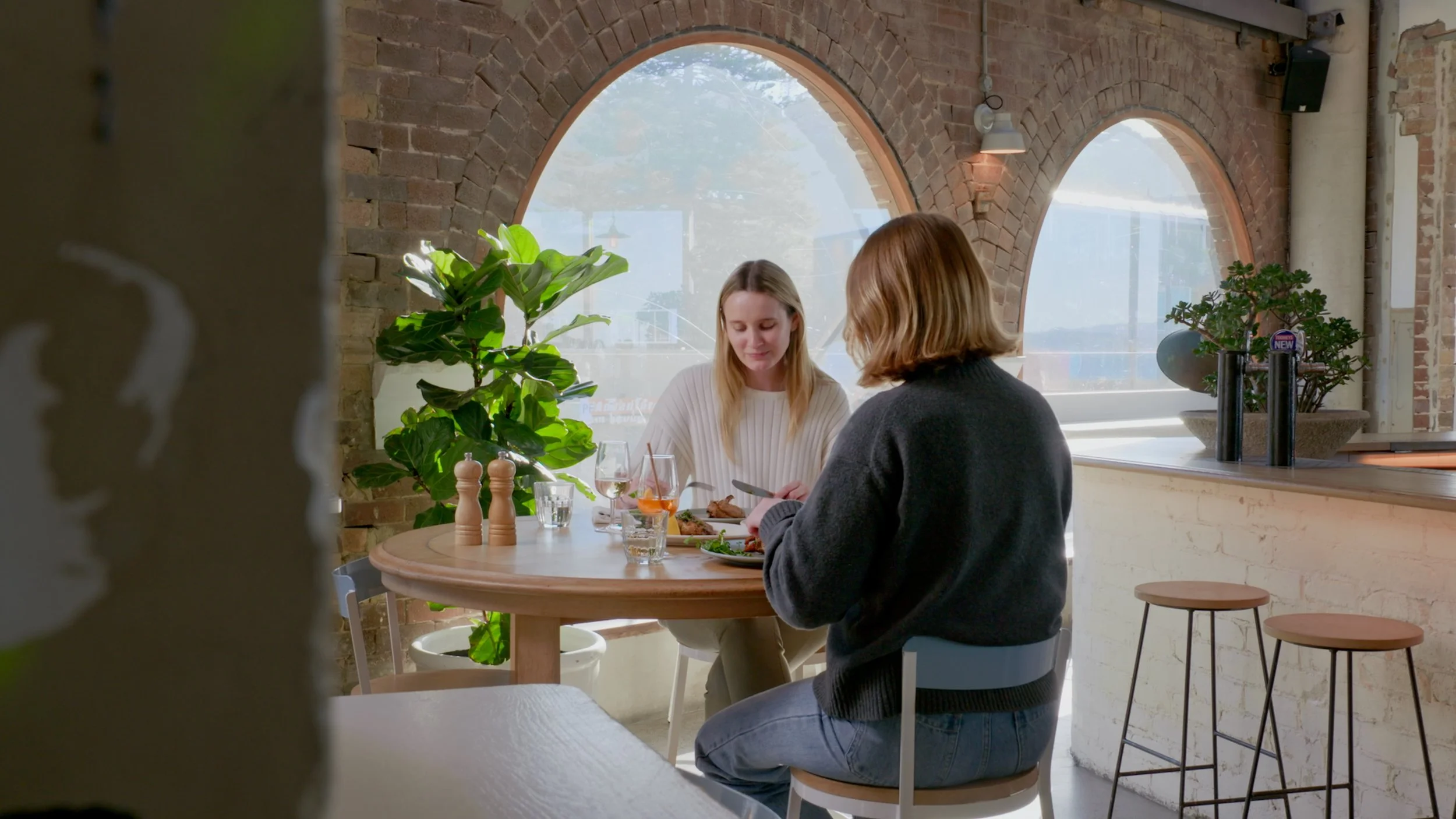 Two women sit at a round dining table in a brick-walled cafe with large arched windows, green plants, and natural lighting, sharing a meal.