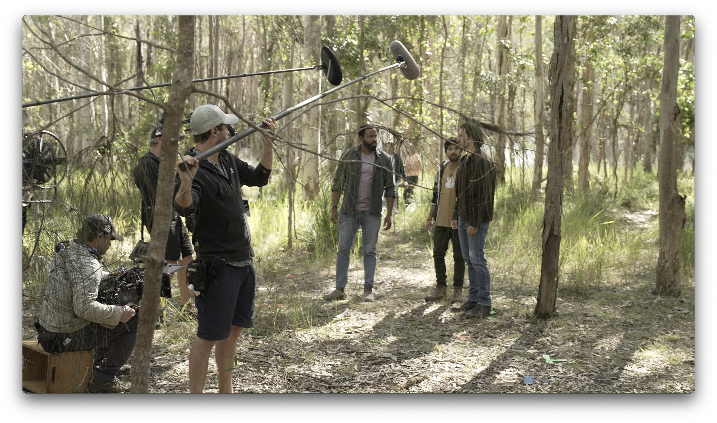 A film crew working in a forest, with one person holding a boom microphone overhead and three others standing and talking among the trees.