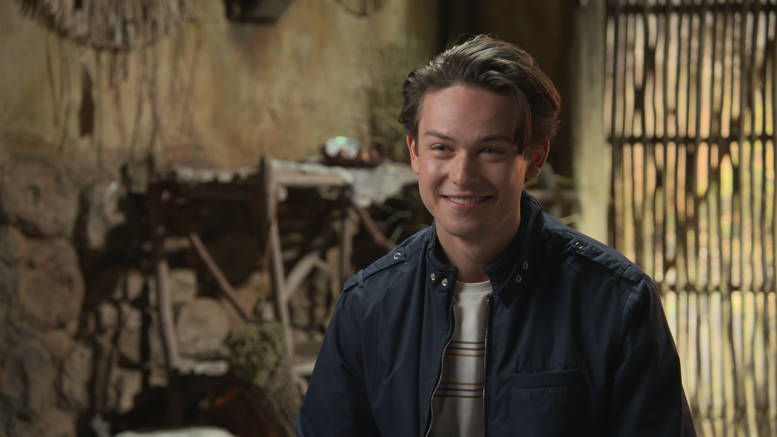 A young man with dark hair smiling, wearing a navy jacket over a white T-shirt with stripes, standing inside a rustic building with wooden walls and a window with vertical slats.