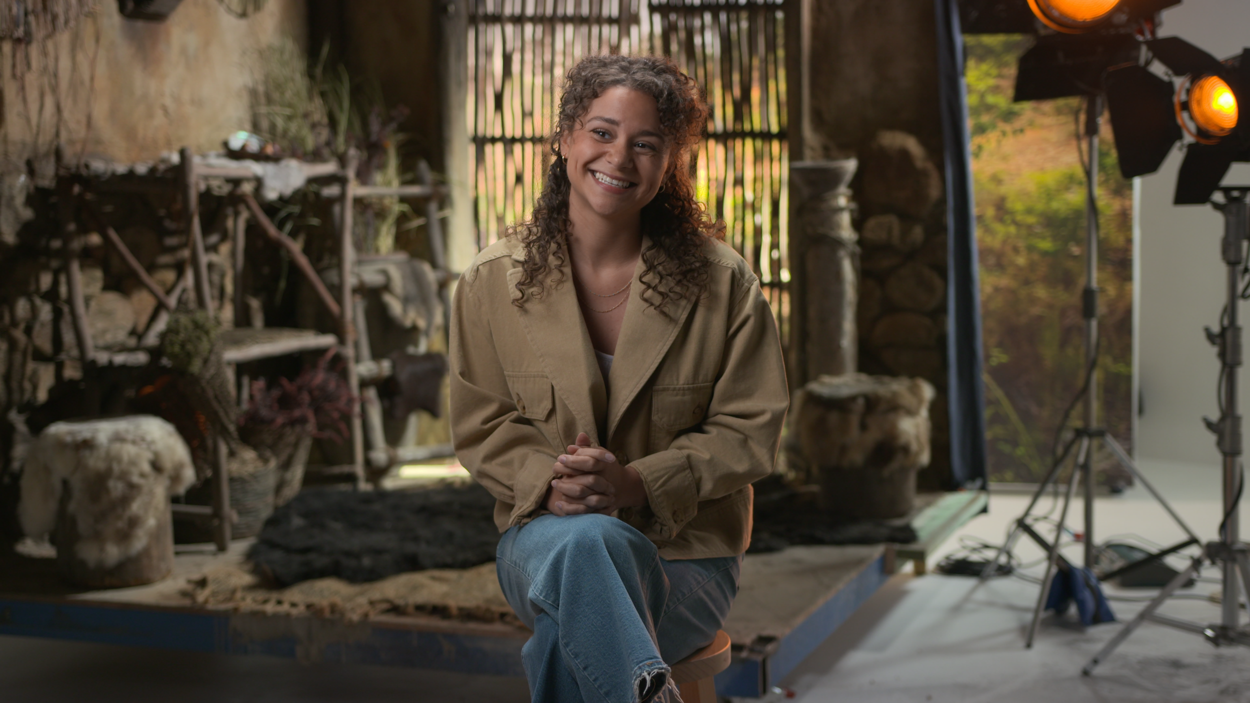 A woman with curly hair smiling with hands clasped, seated in front of a rustic background with wooden shelves, animal pelts, and natural decor, lit by studio lighting.
